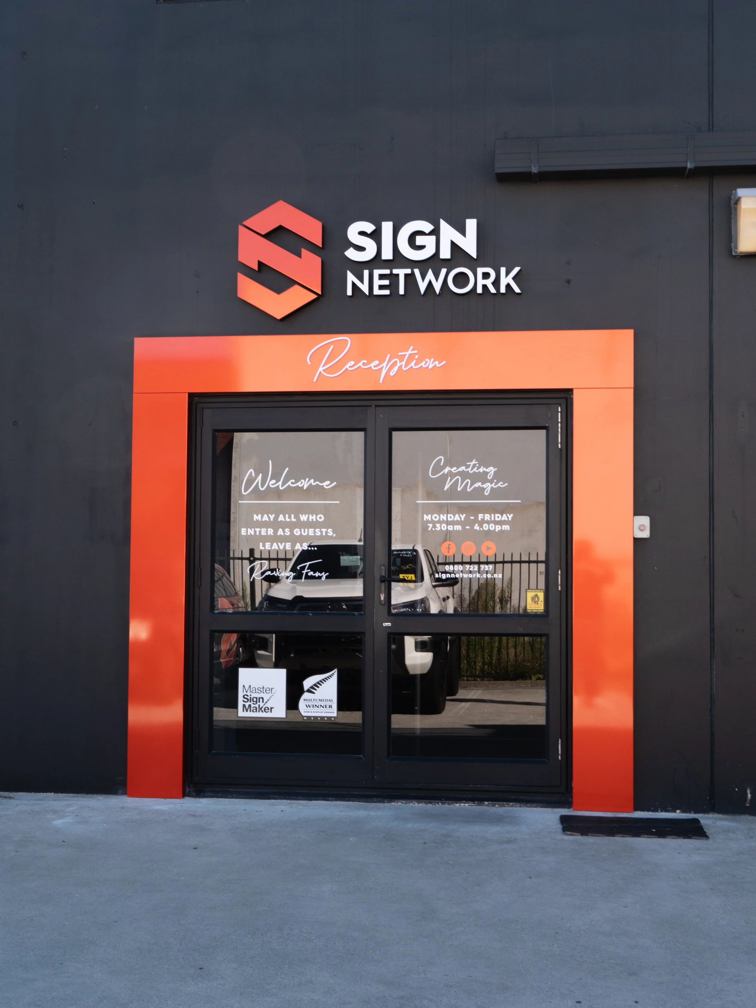 Exterior view of a business called Sign Network with a black building and a bright orange door frame labeled Reception. The sign above the door features a logo with geometric shapes and text that reads 'SIGN NETWORK.' The windows of the door display various signs, including 'Welcome,' 'Creating Magic,' and business hours.