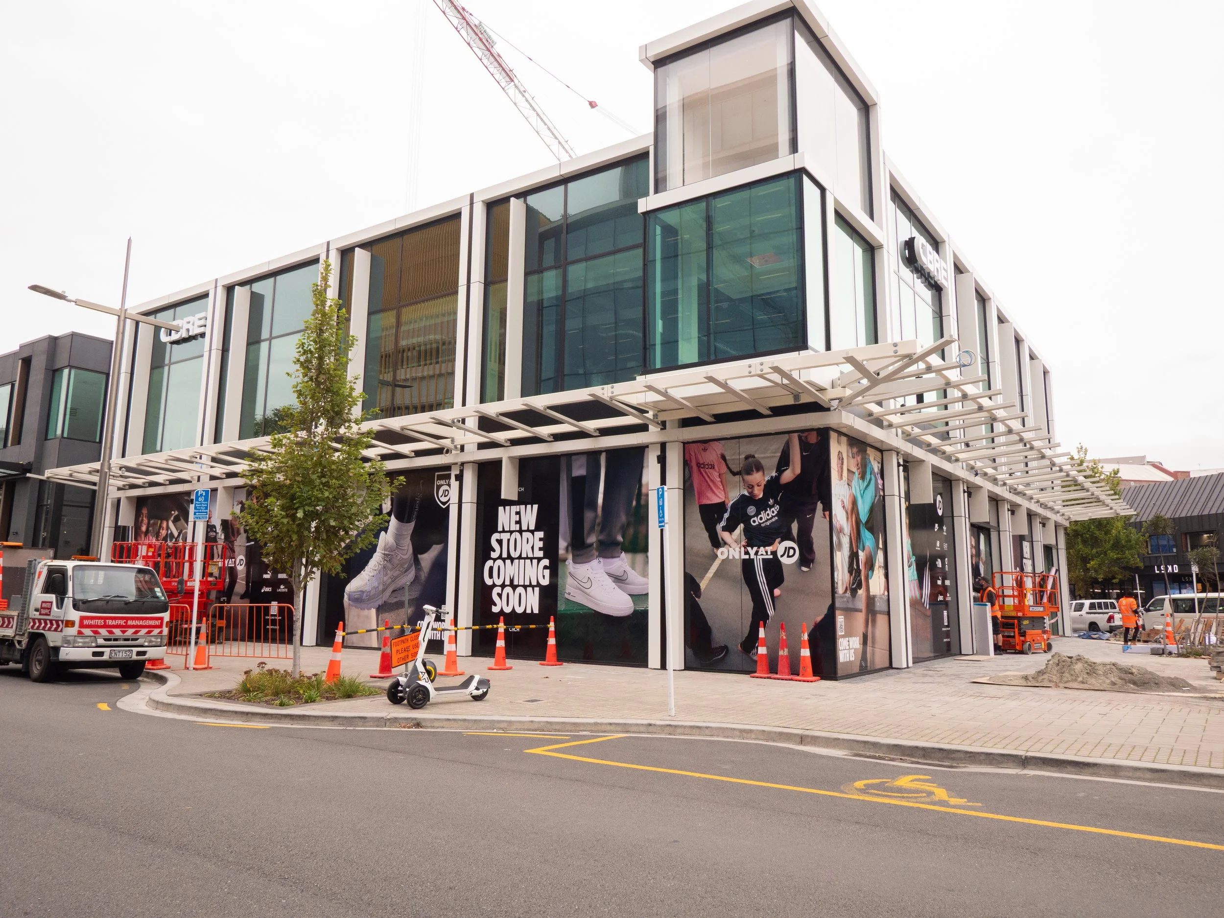 Cleaning and construction workers at a modern shopping center under construction, with signs advertising a new store coming soon, featuring large advertisements of athletic shoes and people exercising.