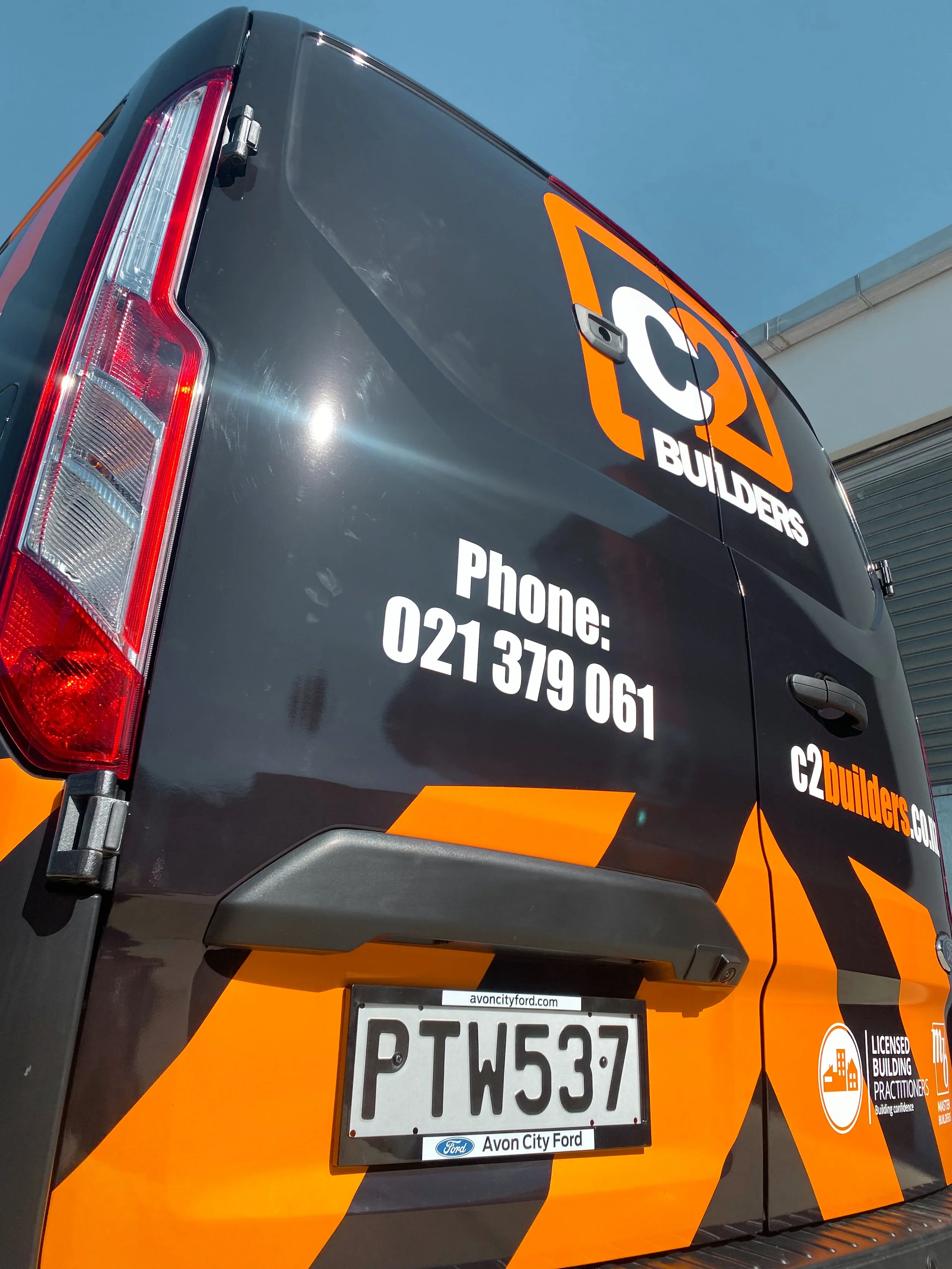 The back of a black and orange utility van with company branding, phone number, website, and license plate, parked outdoors under a clear blue sky.