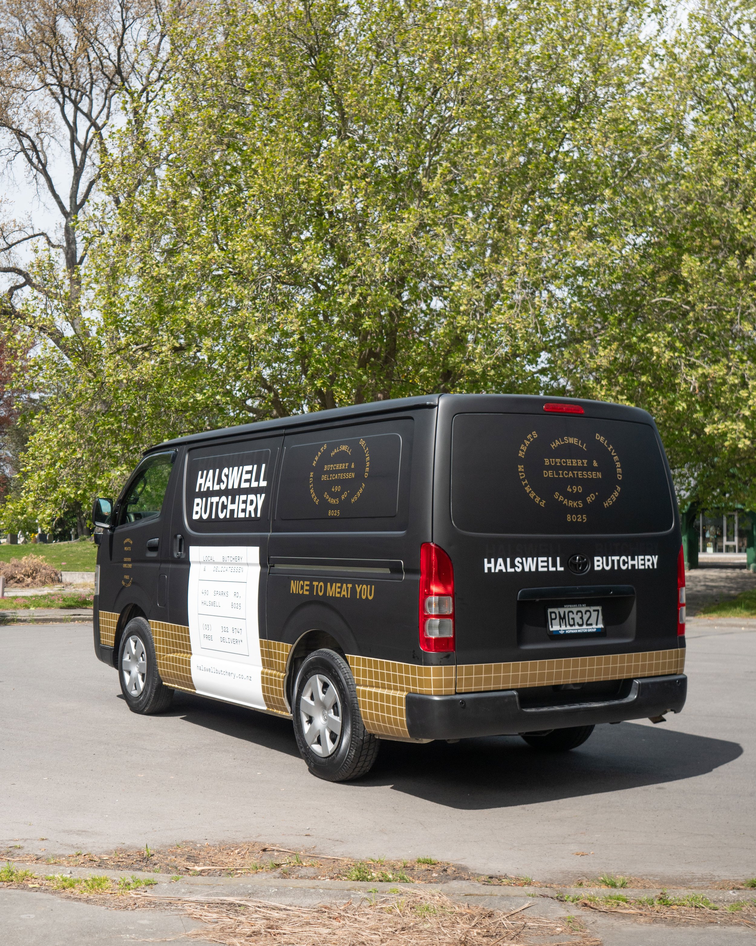 Black delivery van for Halswell Butchery with gold and white text and logo, parked outdoors near trees and a sidewalk.