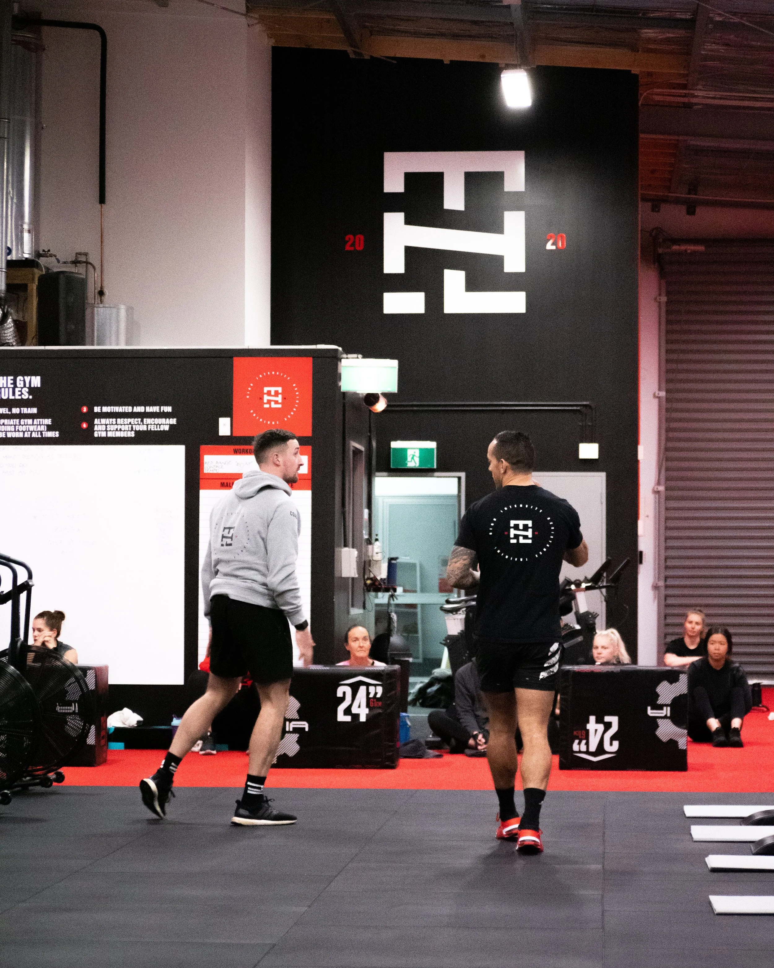 Two men are conversing in a gym with black flooring and red accents. In the background, several people sit on the floor near black boxes labeled 24, 20, and 24. The gym's large logo on the wall reads 'F2' flipped vertically.