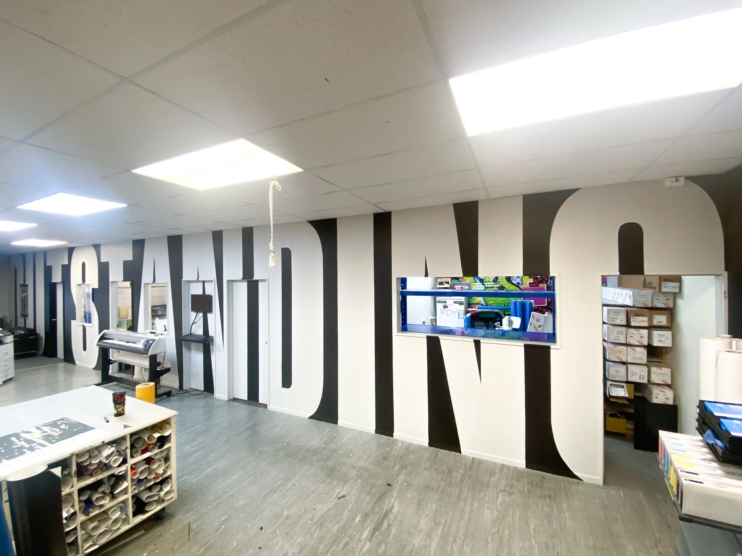 Interior of an office space with white walls featuring black large letters and a window, with shelves, a printer, and office supplies.