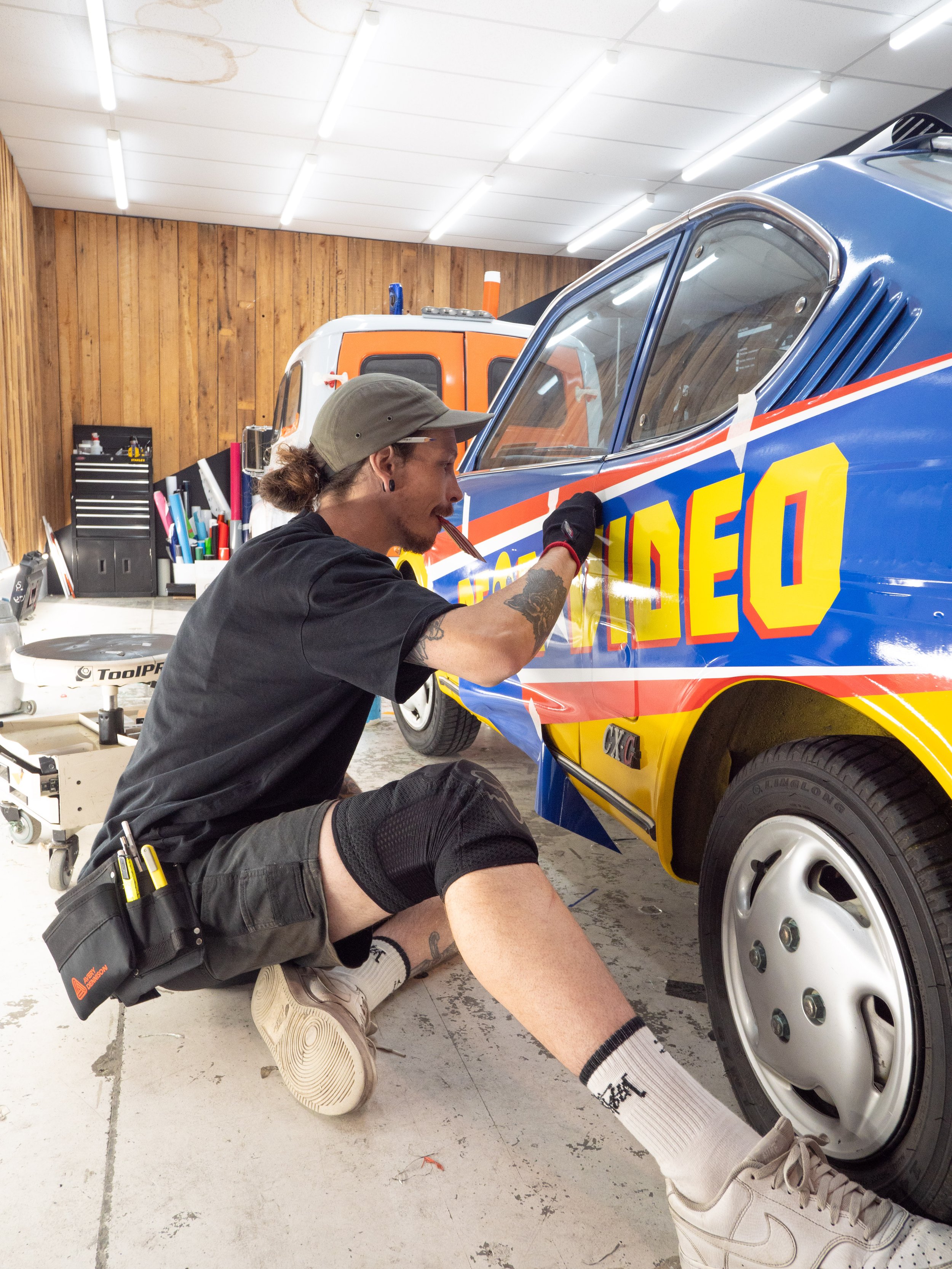 A person with a ponytail, gray cap, black t-shirt, and shorts, painting a colorful race car with the word 'VIDEO' on it inside a garage.