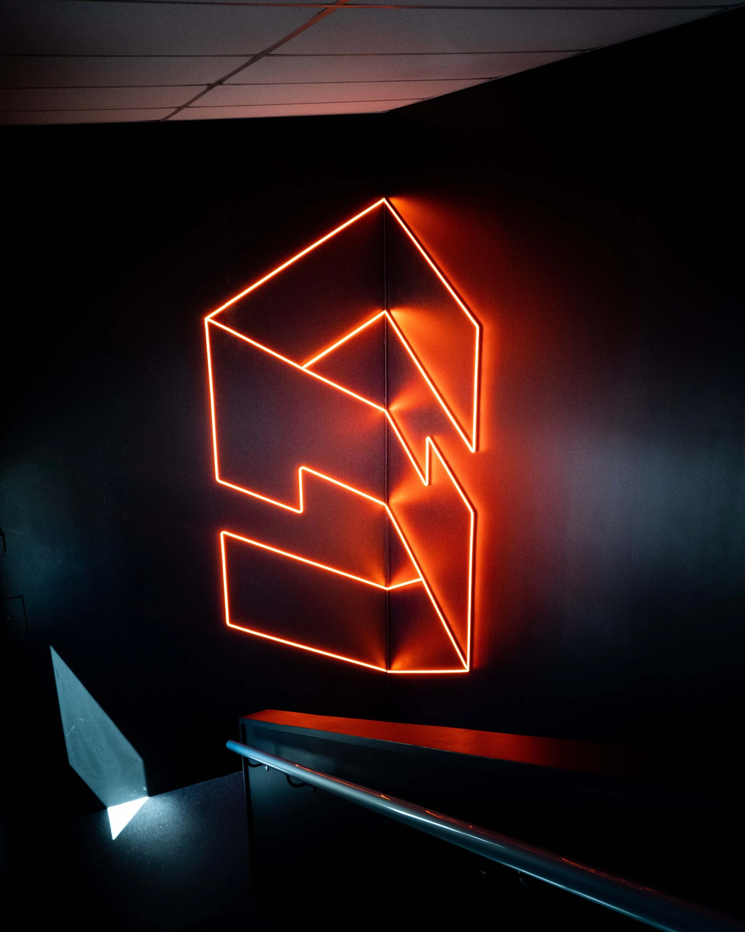 Neon orange geometric art installation on a dark wall, with sections resembling a stylized face or abstract shape, illuminated in a dimly lit room.