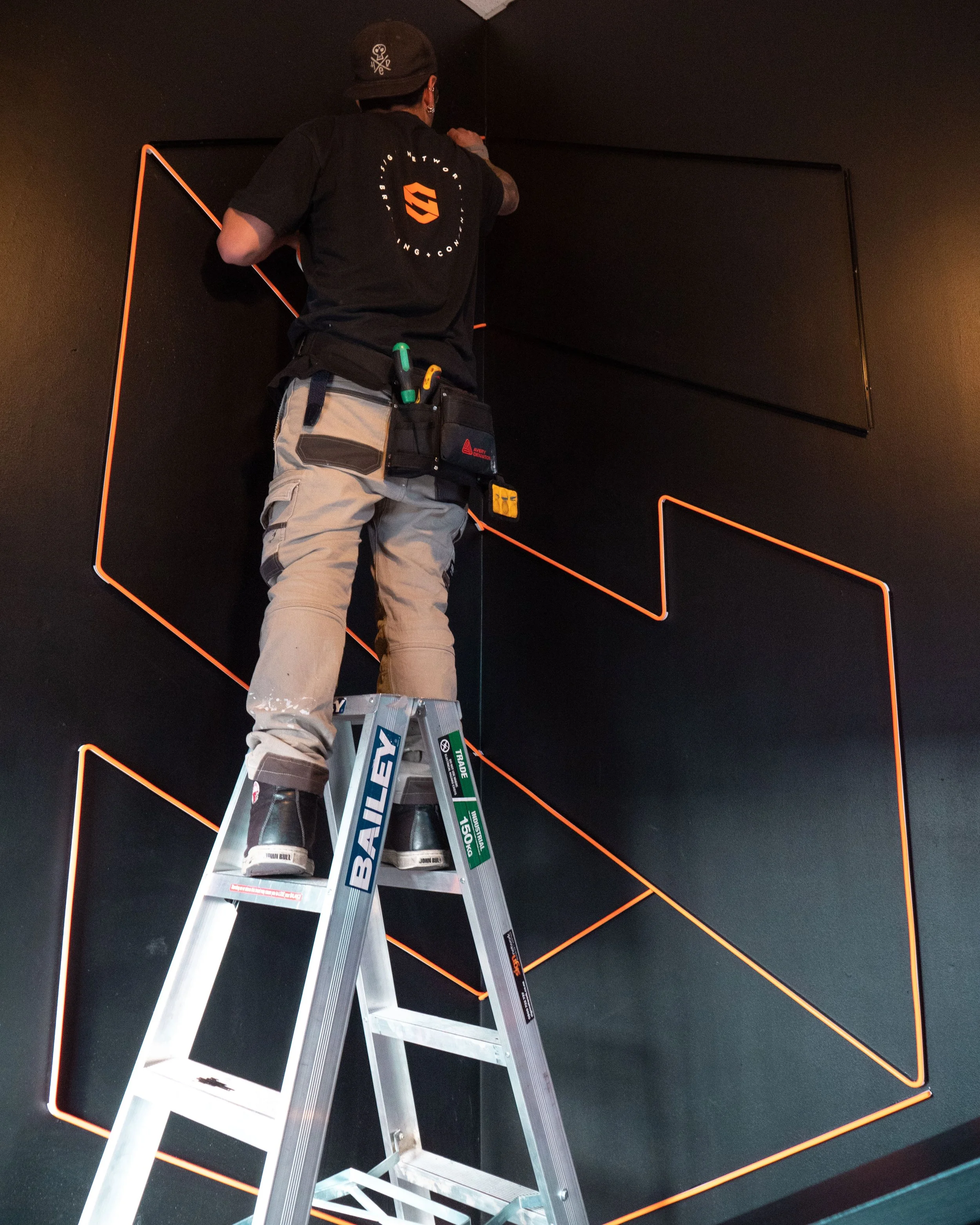 A worker on a ladder installing or repairing neon light strips on a black wall with geometric patterns.