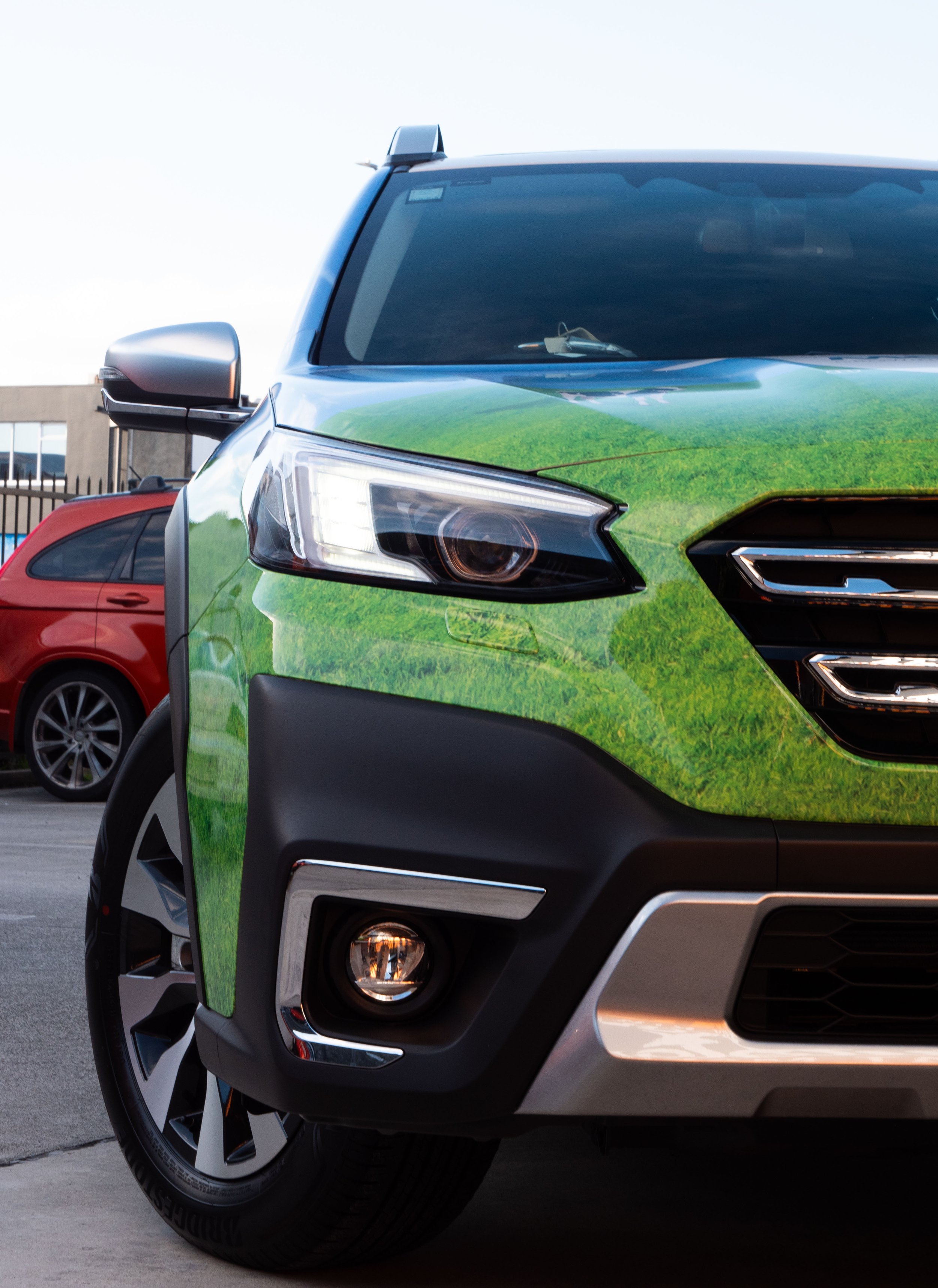 Close-up of the front of a car with a green nature-themed wrap, parked next to a red vehicle in a parking lot.