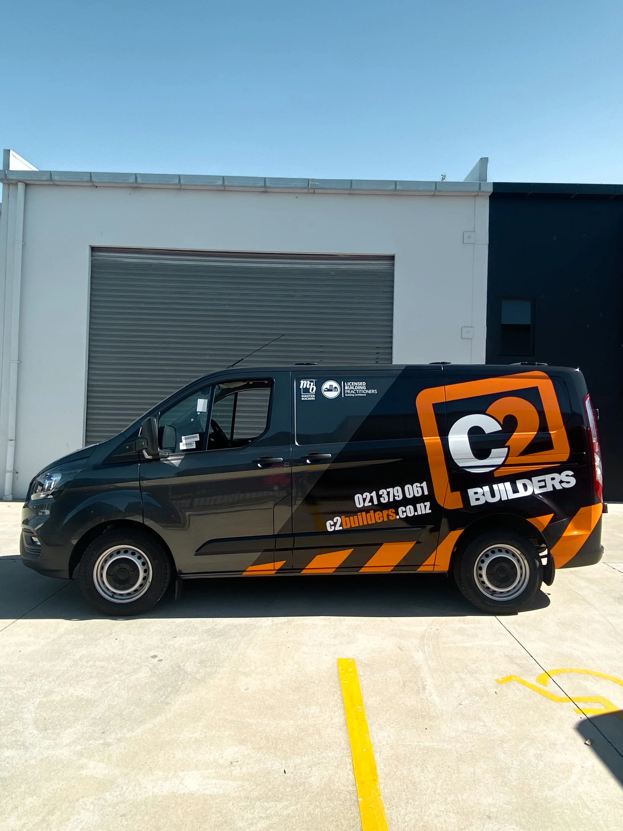 Black C2 Builders branded service van parked in front of a warehouse with gray roller door, blue sky overhead.