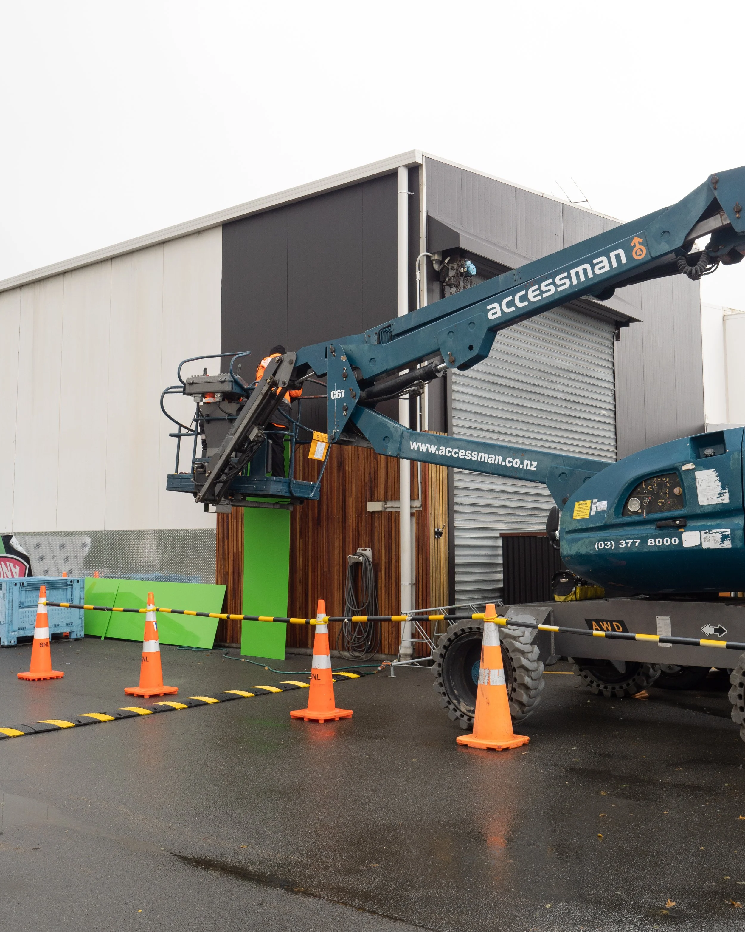 A construction lift with workers installing or maintaining a building facade, surrounded by traffic cones and safety barriers.
