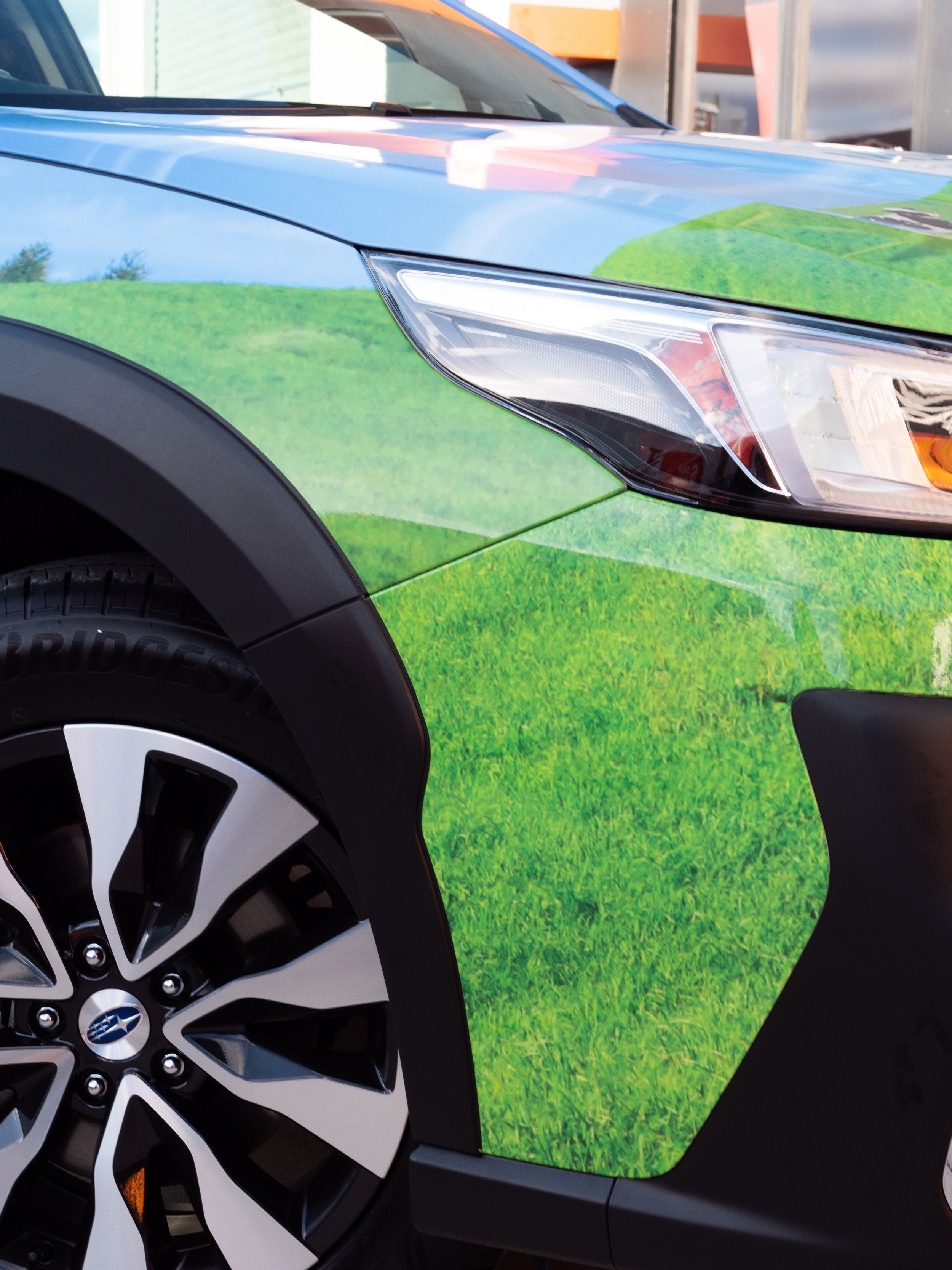 Close-up of the front part of a blue car with a wrap design resembling green grass, showing the headlight, black wheel arch, and part of the tire.