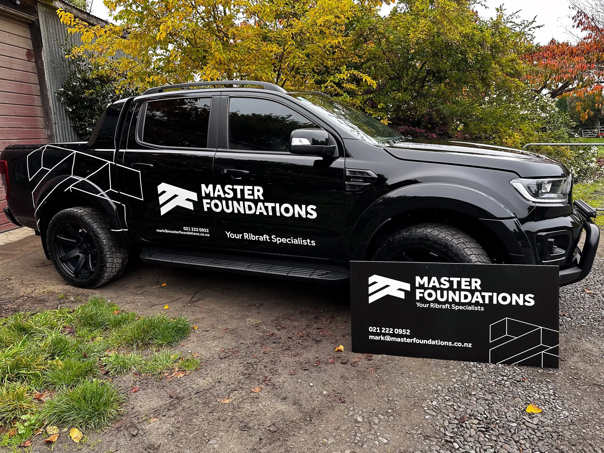 Black pickup truck with company branding for Master Foundations, parked outdoors near trees with yellow and red leaves, and a promotional sign on the ground with the company's logo, contact information, and slogan.