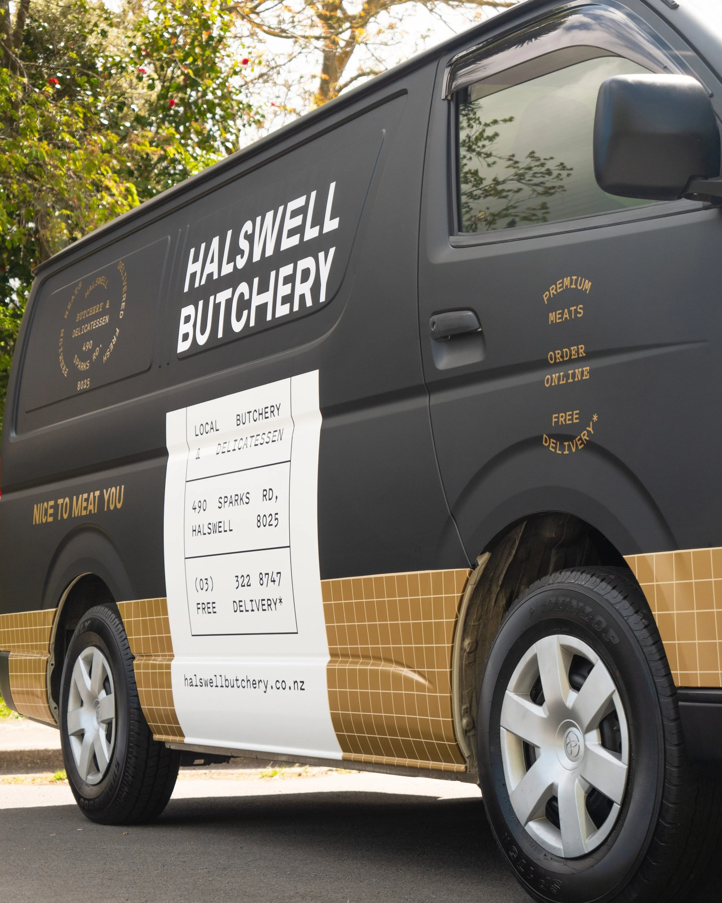 Black van with yellow and white signage for Halswell Butchery, a local butcher and delicatessen, featuring contact information, website, and advertisements for online ordering and free delivery.