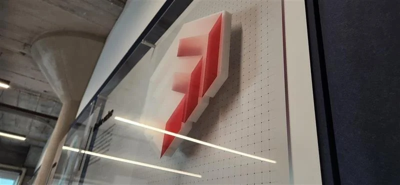 A framed piece of modern art featuring a three-dimensional red and white geometric shape on a white dotted background, displayed on a wall behind glass.