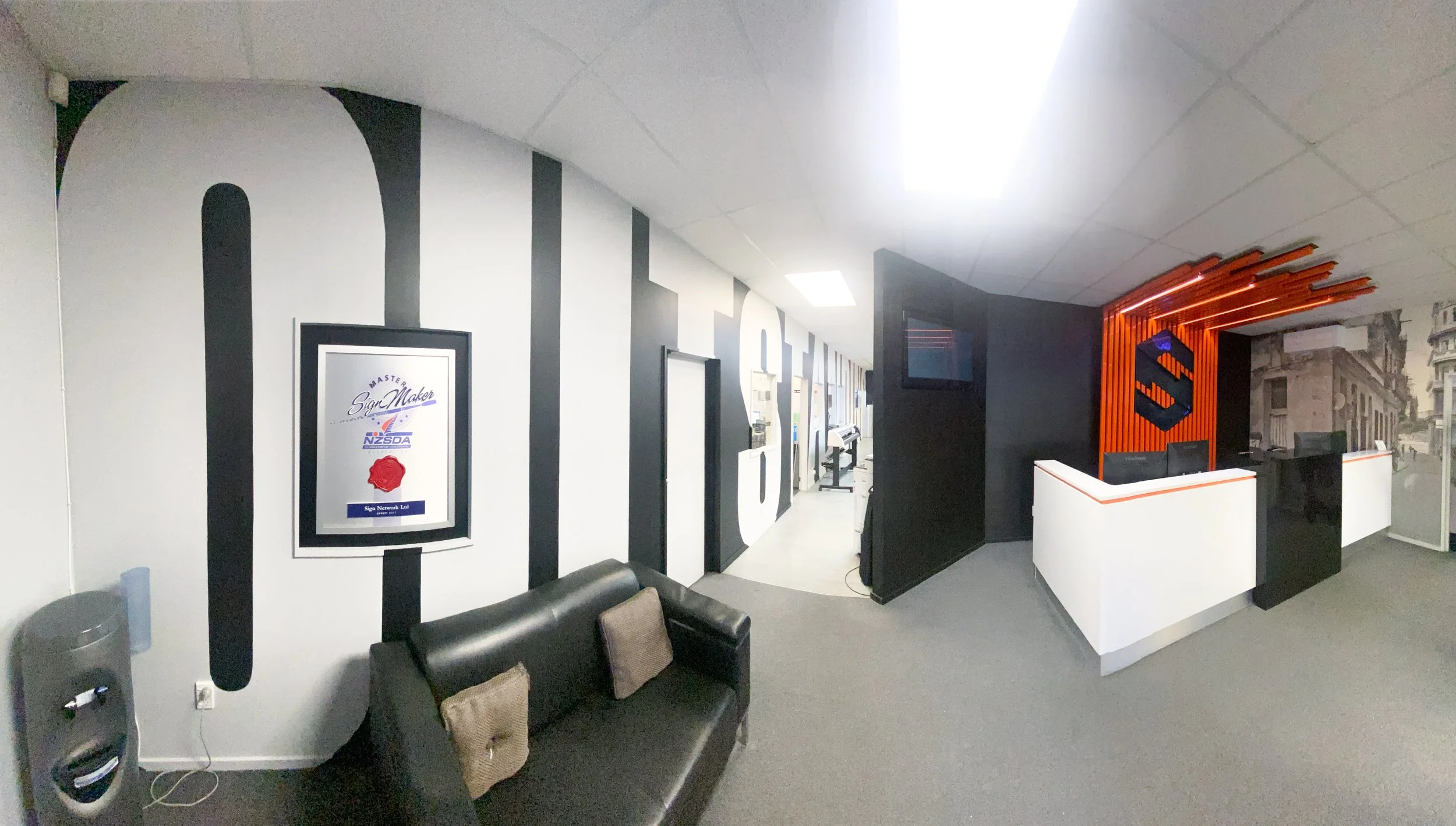Office reception area with a black and white color scheme, a black sofa with two brown pillows, and a sign with a red wax seal, logo, and text on the wall. The reception desk has black, white, and orange accents, with orange slats and a black logo. There is a grayscale cityscape mural on the wall behind the desk.