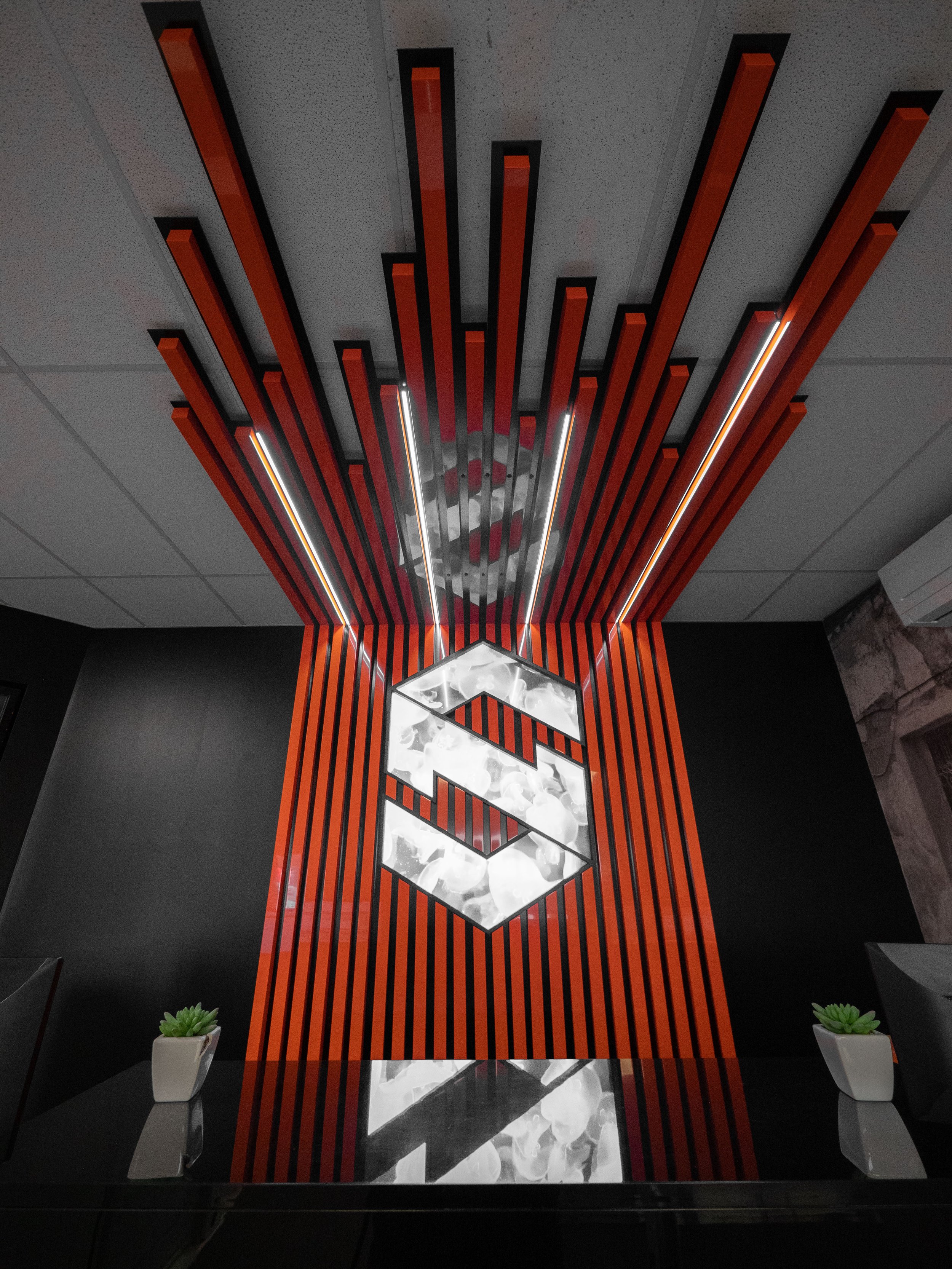 Interior ceiling with modern red and black wooden slats, illuminated geometric hanging light fixture with a hexagonal shape, black walls, and small plants on a table.