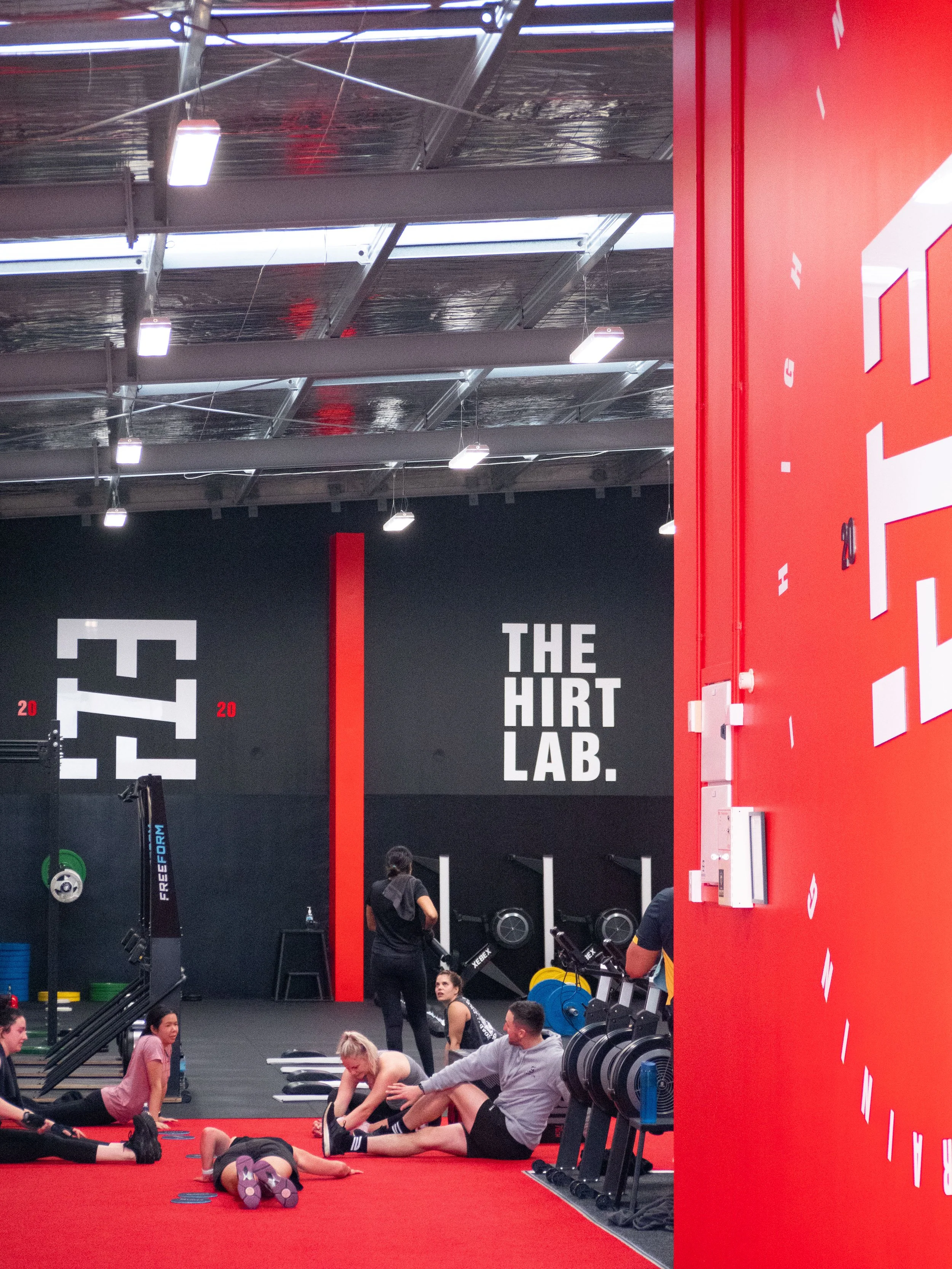 People exercising at The Hirt Lab gym, with workout equipment and a large red wall and black wall with white text in the background.