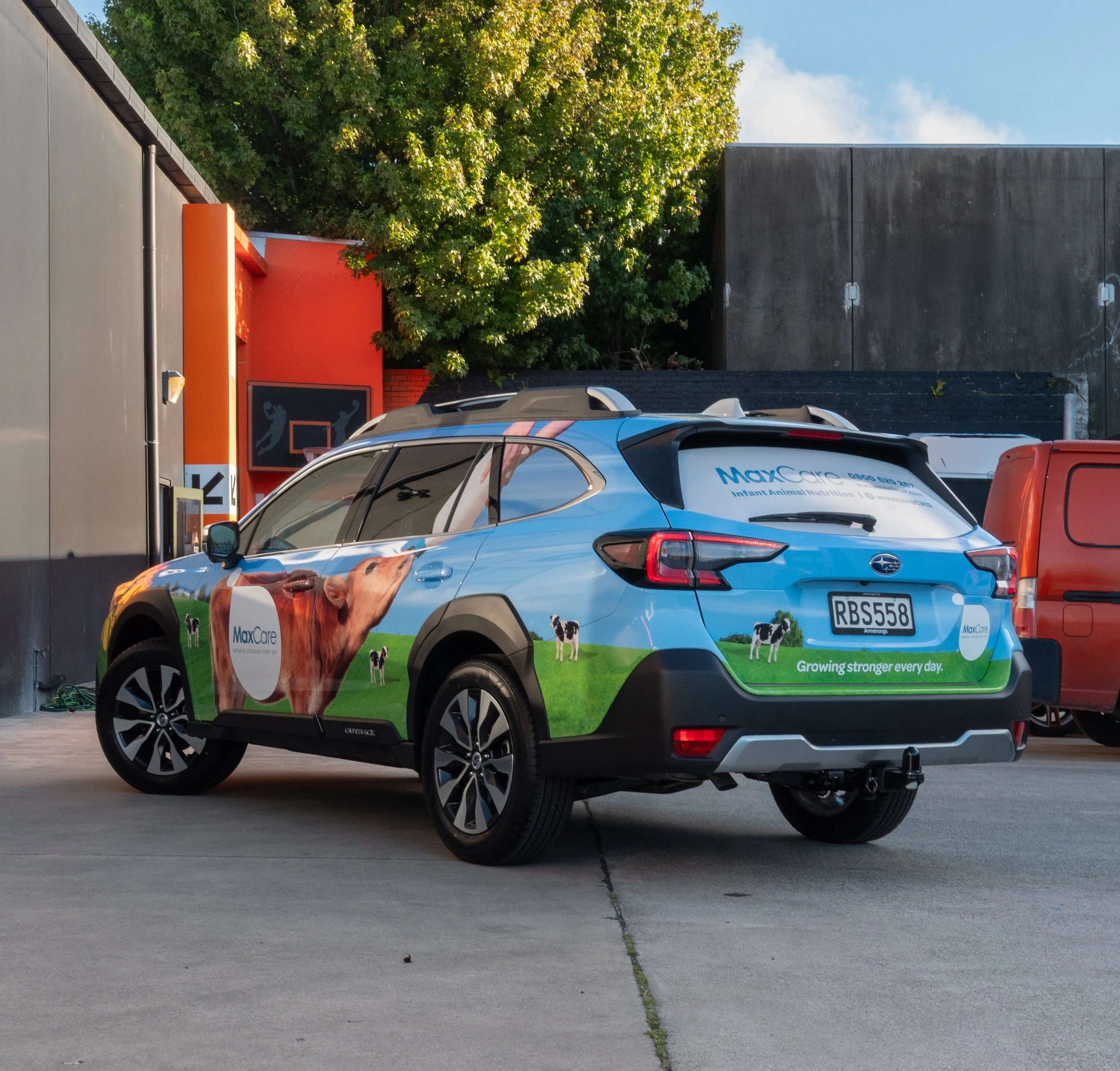 A car with MaxCare branding featuring images of cows and a green field, parked in an outdoor lot.