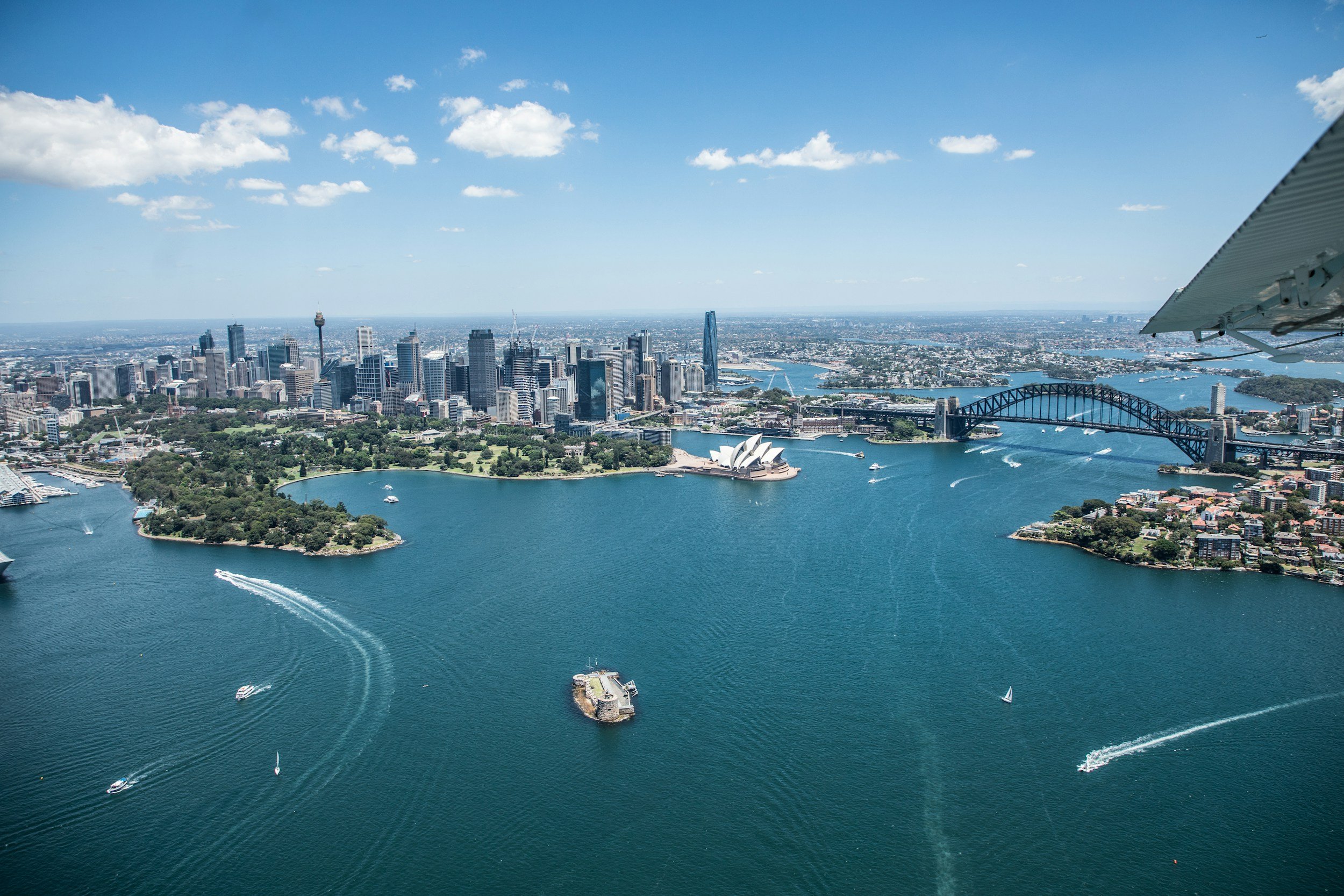 Aerial view of Sydney Harbor with the Sydney Opera House in the center and Sydney Harbour Bridge on the right. The city skyline is visible in the background under a clear blue sky.