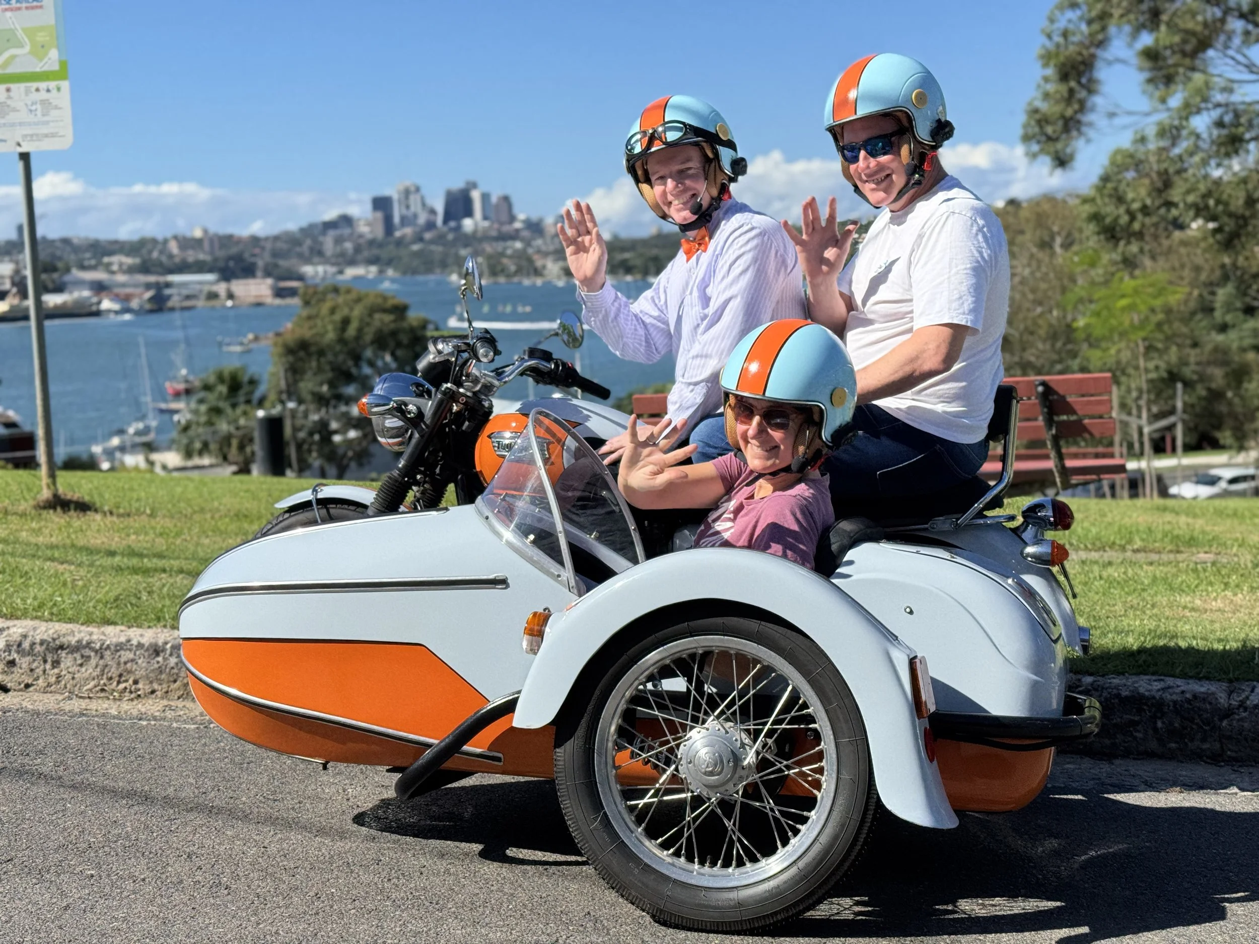 Three people wearing helmets on a motorcycle with a sidecar, parked by a waterfront with city skyline and trees in the background.