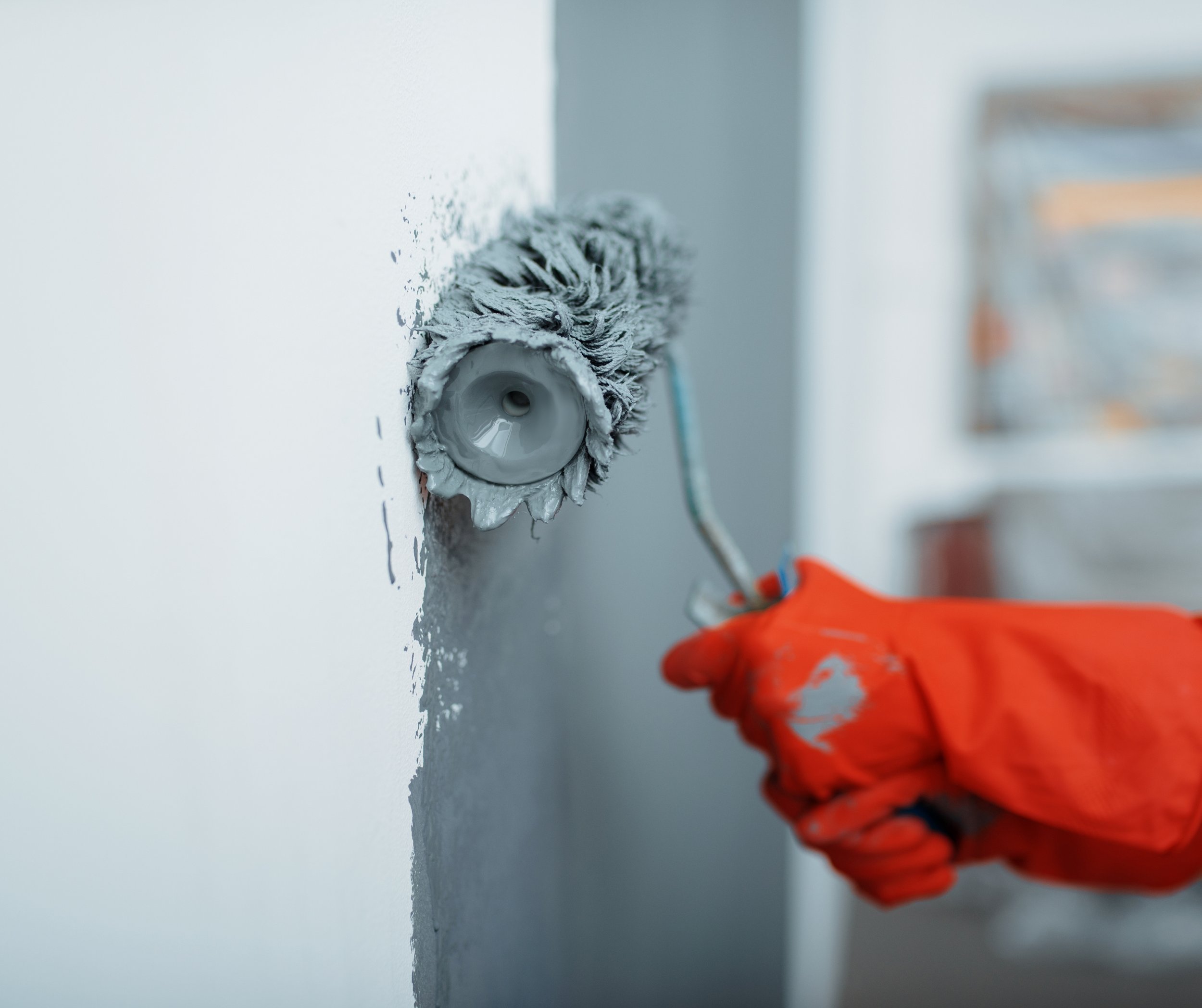 Close-up of a paint roller with gray paint, held by a person wearing a bright orange glove, applying paint on a white wall.