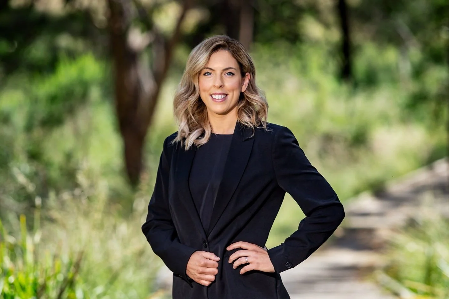 A woman with shoulder-length blonde hair and a black blazer standing outdoors in a green park or forest, smiling.