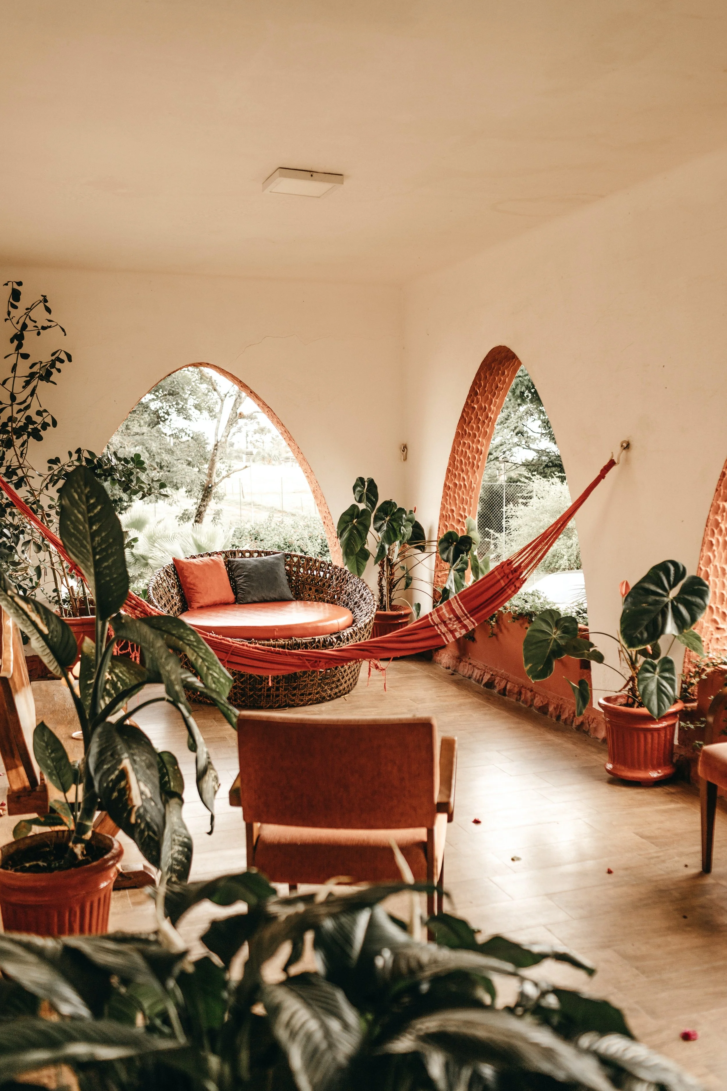 A cozy porch area with a circular wicker loveseat with black and orange pillows, surrounded by green plants, arched windows, and a hammock, with trees visible outside.