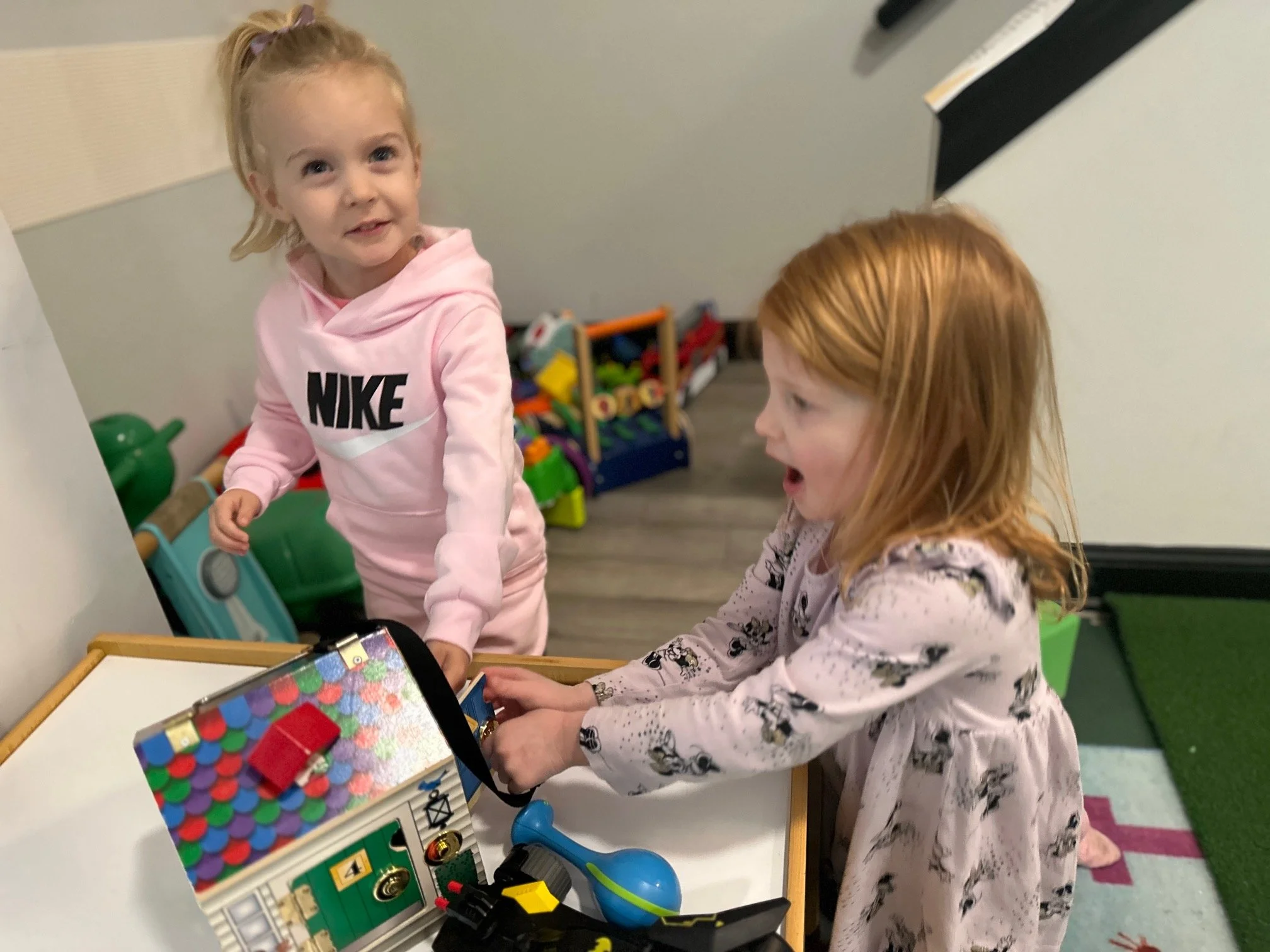 Two young girls playing with a toy cash register on a table in a playroom. One girl with blonde hair and a pink Nike hoodie is standing, while the other girl with red hair and a floral dress is leaning over, pressing buttons.