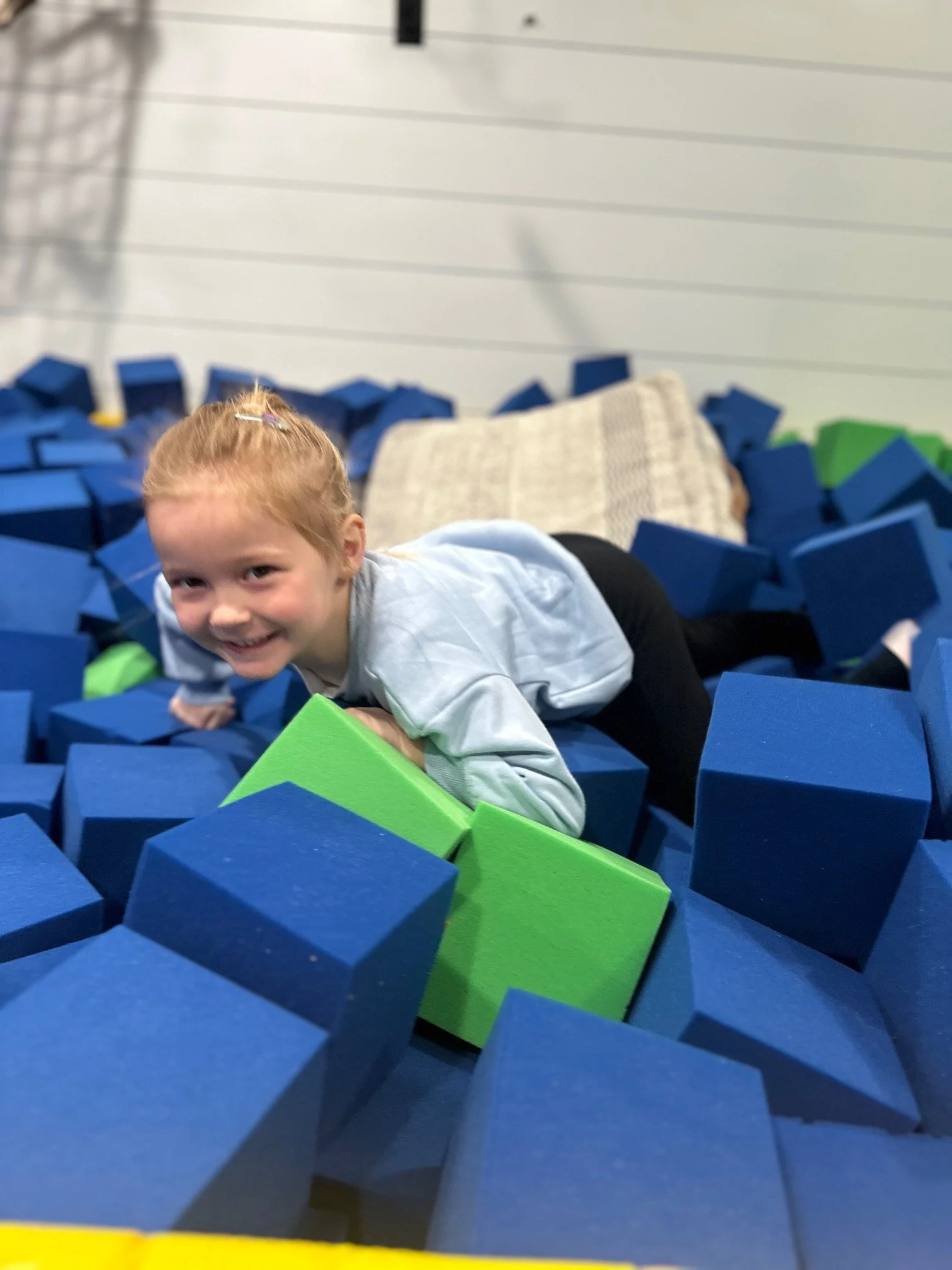 A young girl with red hair and a hair clip smiling while crawling through a foam pit filled with blue, green, and yellow foam blocks.
