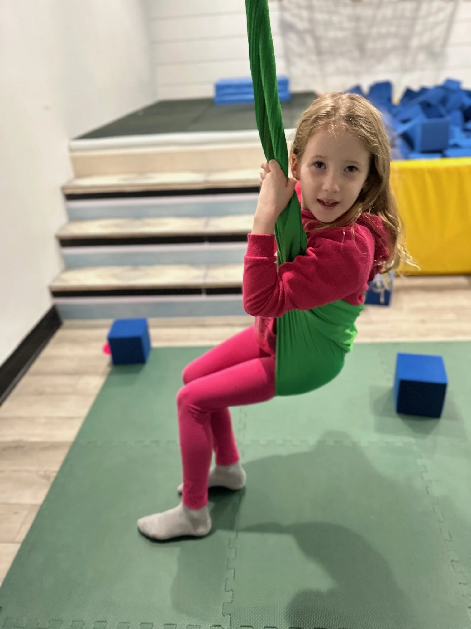 A young girl with red hair in a gym or playroom, sitting on a green mat, holding a green hanging gym ring, with blue foam blocks and a yellow bin in the background.
