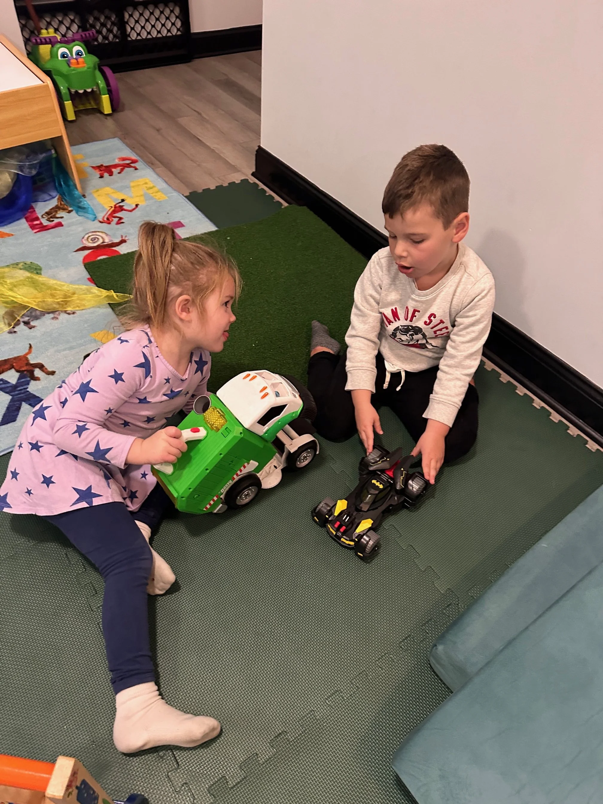 Two children, a girl and a boy, playing with toy cars on a green foam mat in a playroom. The girl is holding a green and white toy dump truck, and the boy is holding a black and yellow race car. The room has colorful toys and a children's rug with an