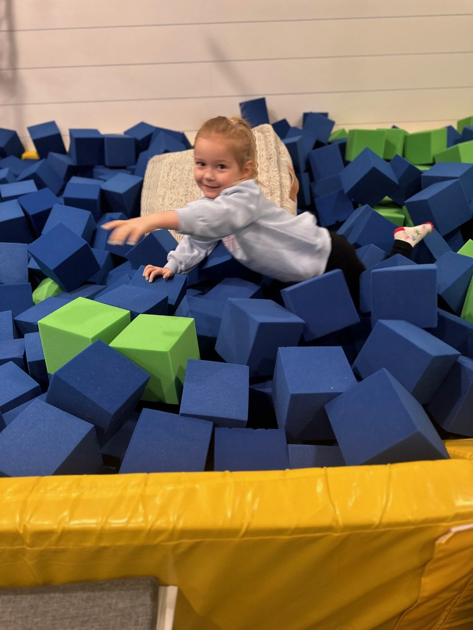 A young girl playing in a foam pit filled with blue, green, and yellow foam blocks at an indoor play area.