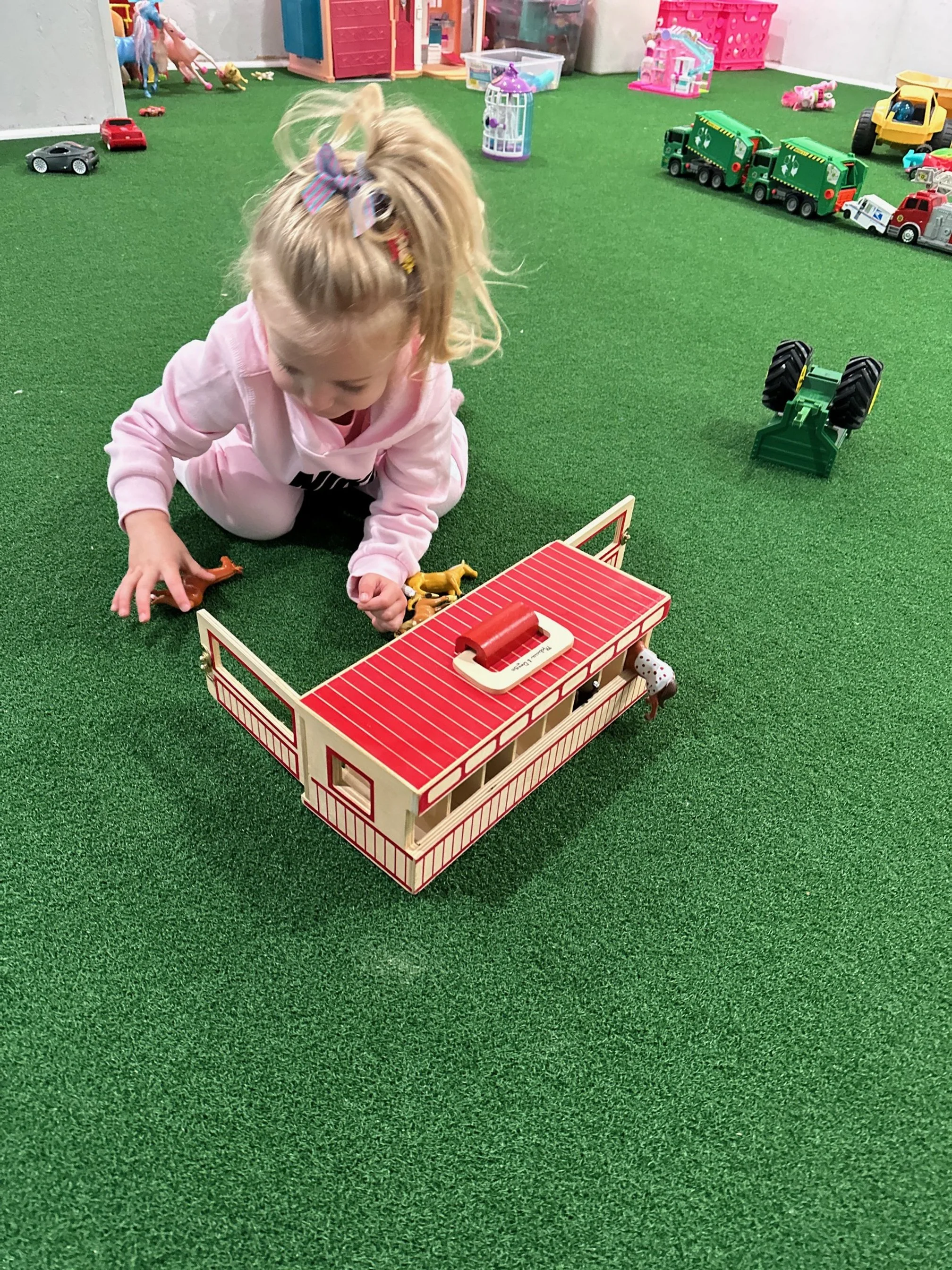 A young girl with blonde hair and a colorful bow on her head, playing with toy animals and a wooden barn on a green indoor play carpet. There are toy cars, trucks, and various colorful toys scattered around.