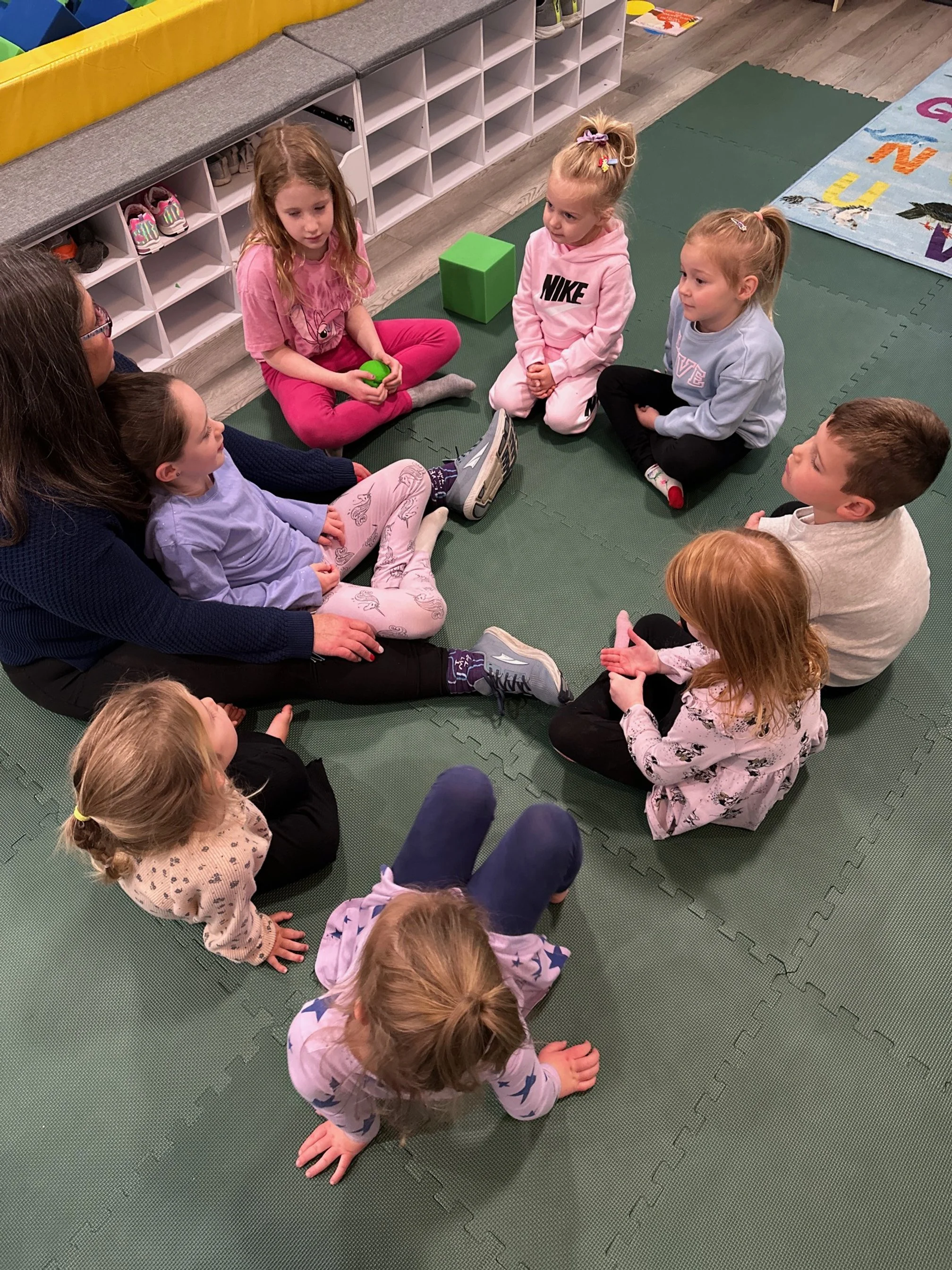 A group of children and an adult sitting in a circle on a green mat, engaging in a group activity in a classroom setting.