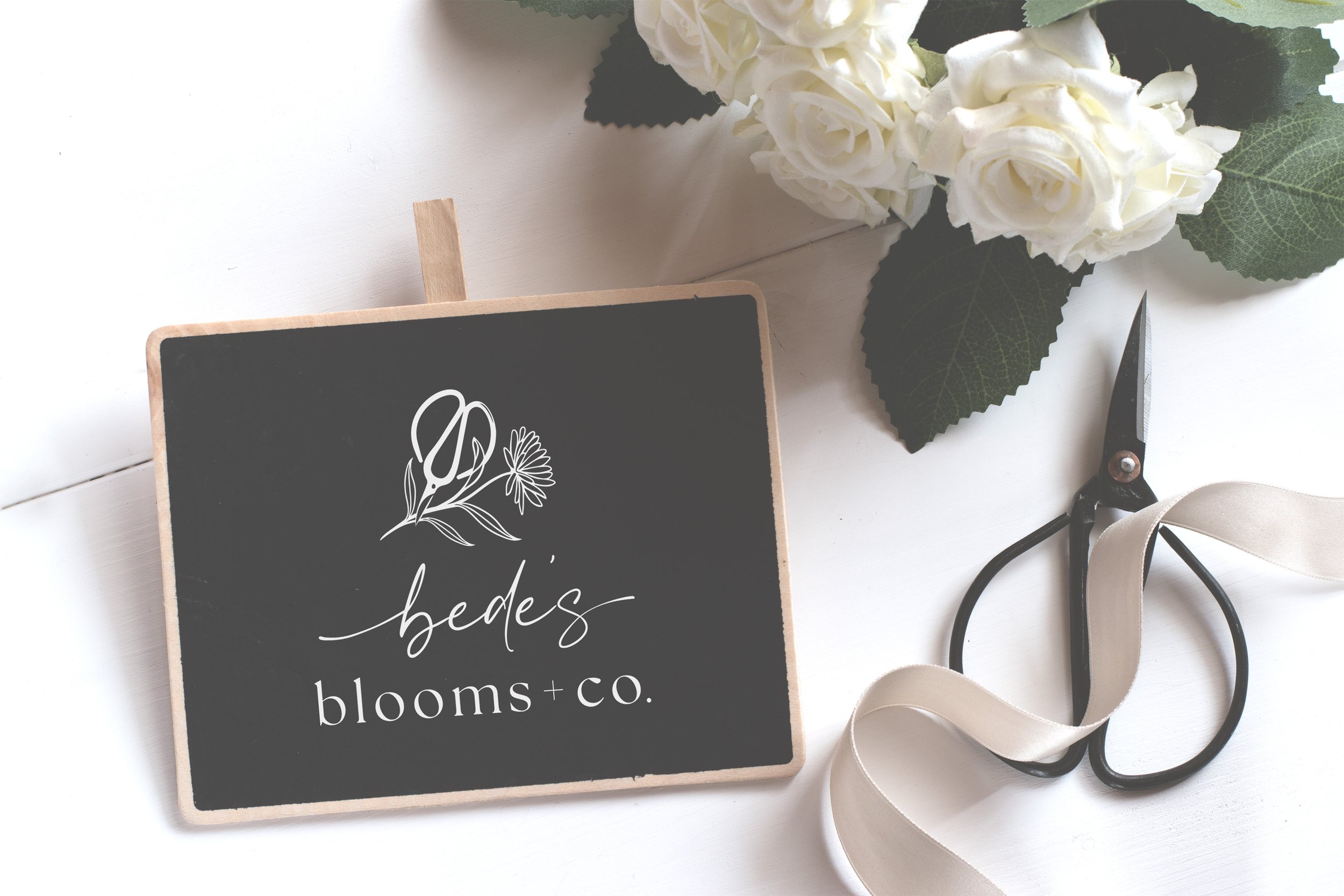 Chalkboard with floral logo reading 'Bede's Blooms + Co.' next to white roses, black scissors, and beige ribbon on a white surface.