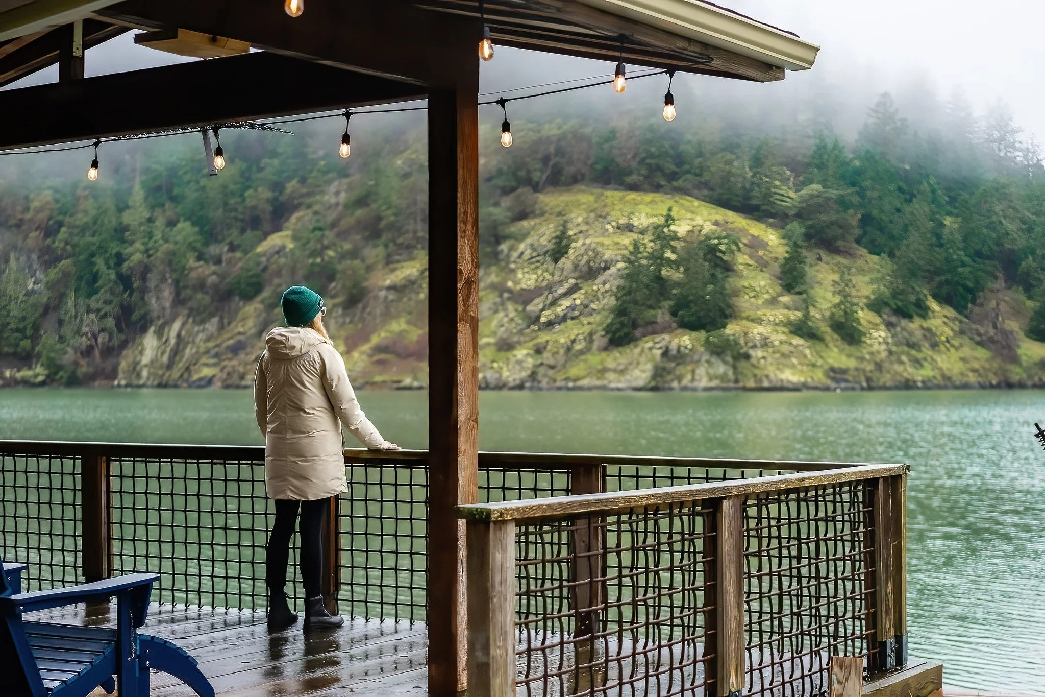 Person standing on a wooden deck overlooking a misty lake with green hills in the background, wearing a beige coat and green beanie, with string lights overhead.
