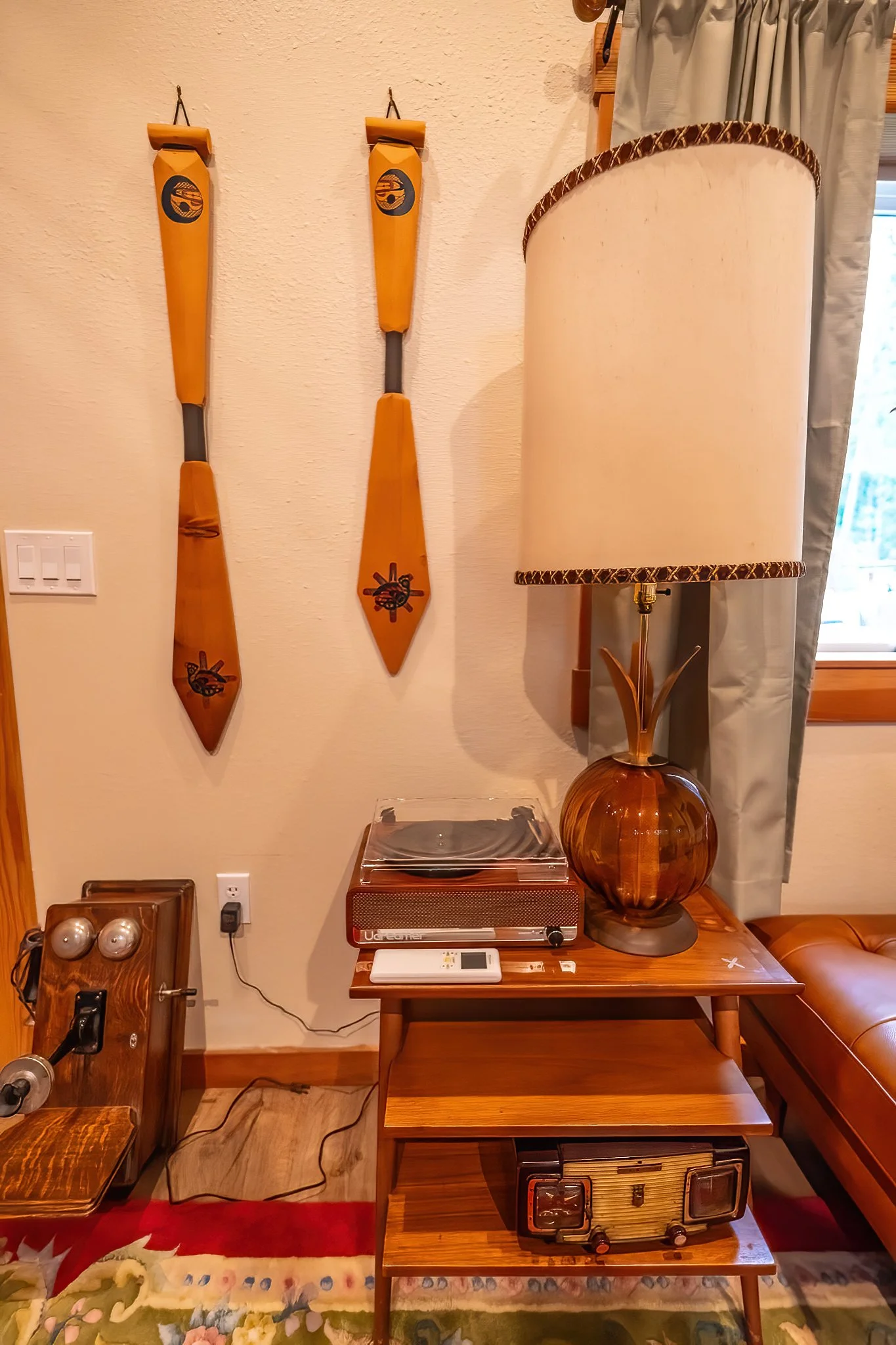 Wall with two wooden paddles hanging, side table with vintage turntable and lamp, and part of a window with curtain.