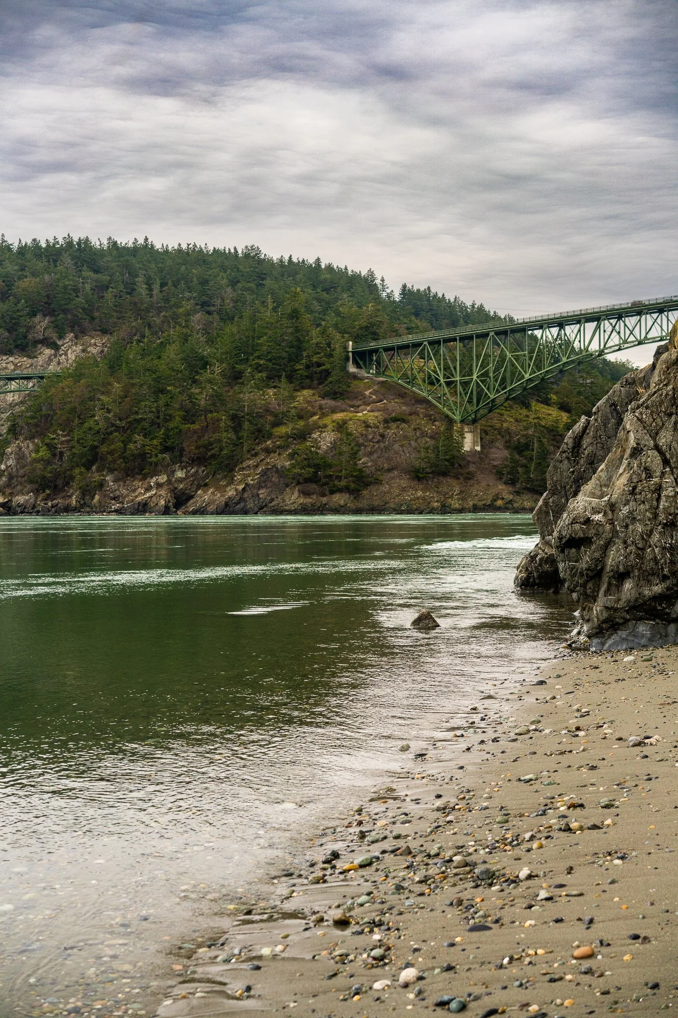 A scenic view of a river with a sandy and pebbly shore on the right, a rocky formation on the right side, and a bridge over a forested hill in the background, under a cloudy sky.