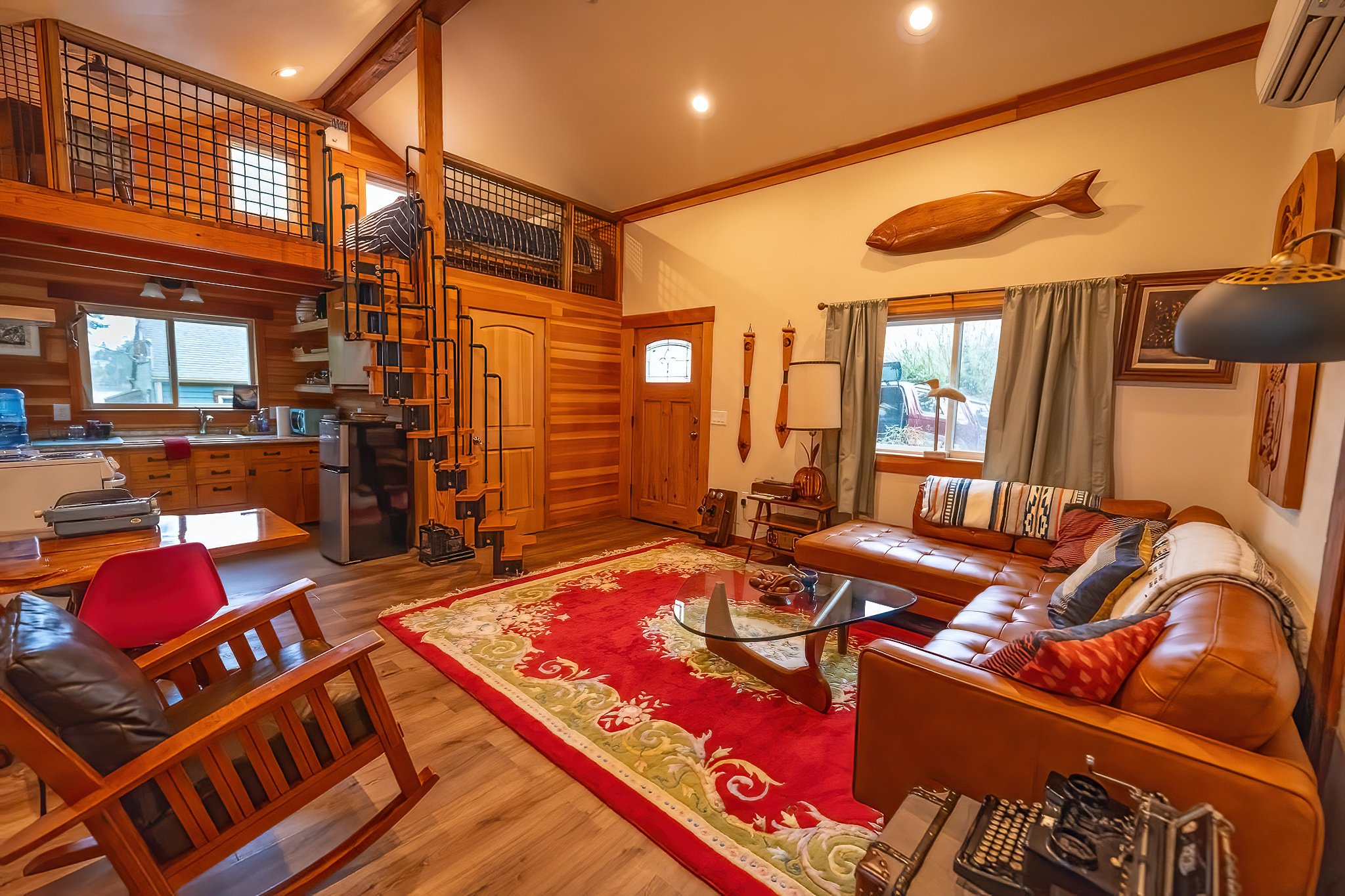 Living room with leather sectional sofa, glass coffee table, area rug, and wooden accents, featuring a wall-mounted wooden fish decor and a spiral staircase leading to a loft area with bed.
