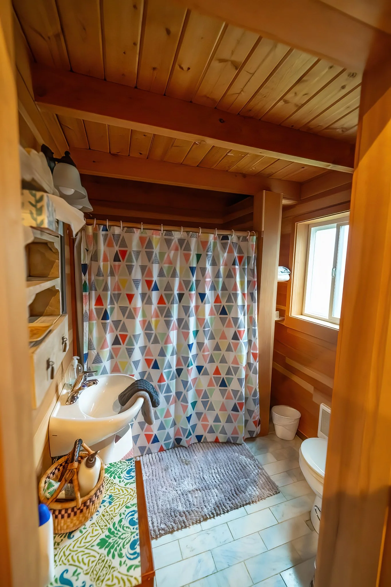 A cozy bathroom with wooden walls and ceiling, a small window, a shower with colorful triangular-patterned curtain, a white sink with a towel, a gray bath mat, and a toilet.