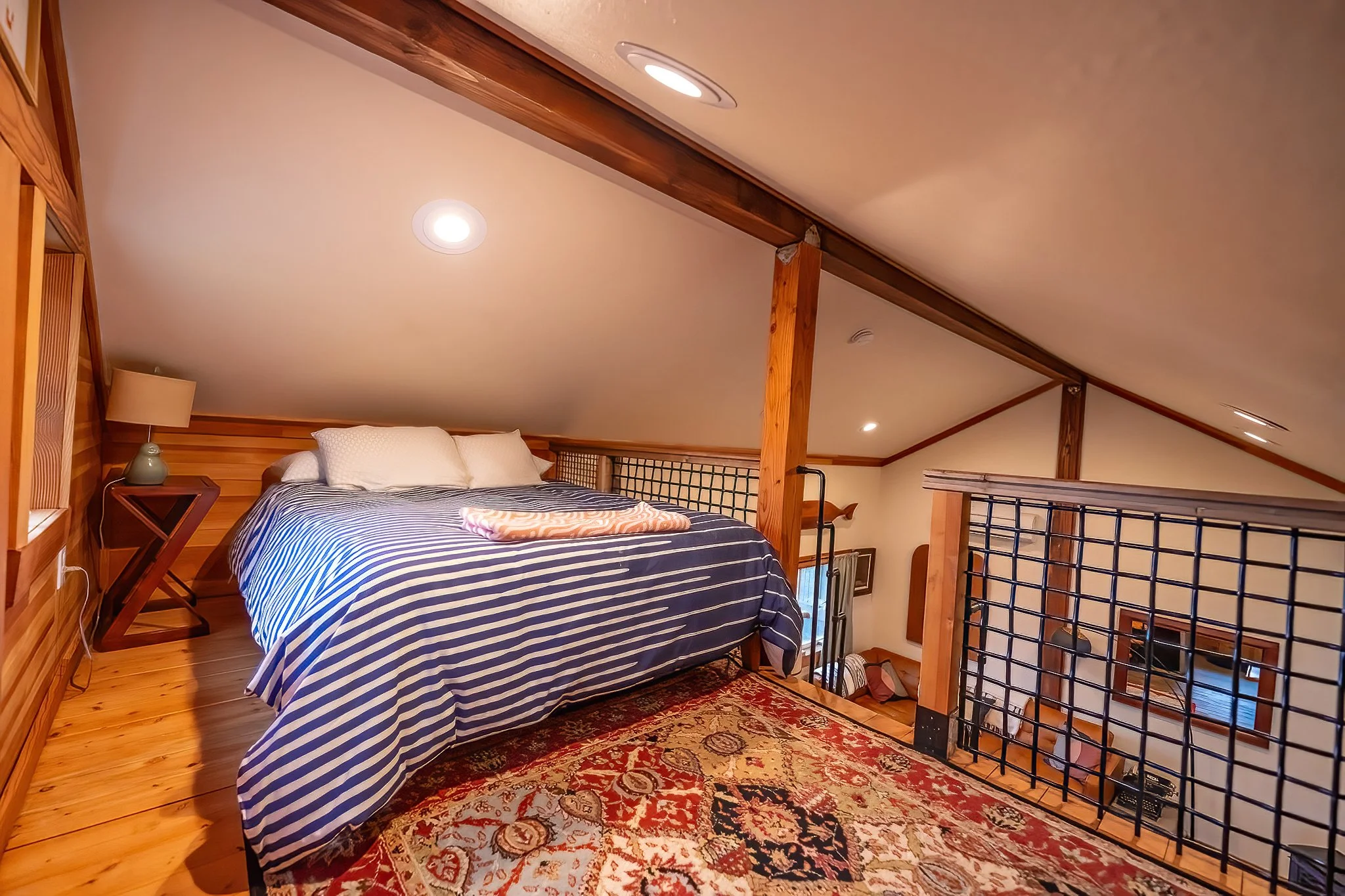Loft bedroom with a bed featuring navy blue and white striped bedding, two pillows, a side table with a lamp, and a colorful patterned rug on a wooden floor. The room has sloped ceilings with recessed lighting and wooden beams.
