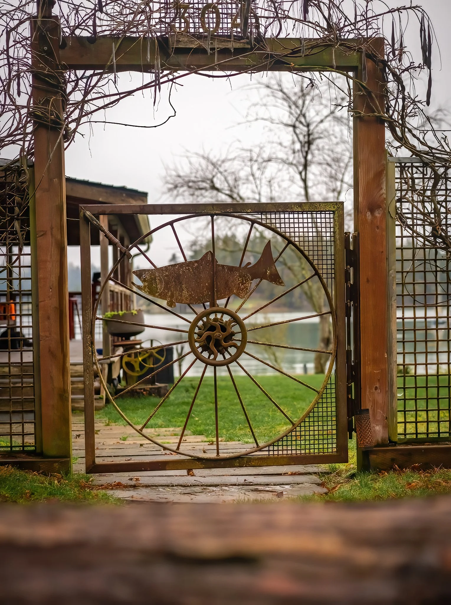 Rusty metal gate with a fish-shaped decoration, opening to a porch overlooking a lake, with trees and cloudy sky in the background.