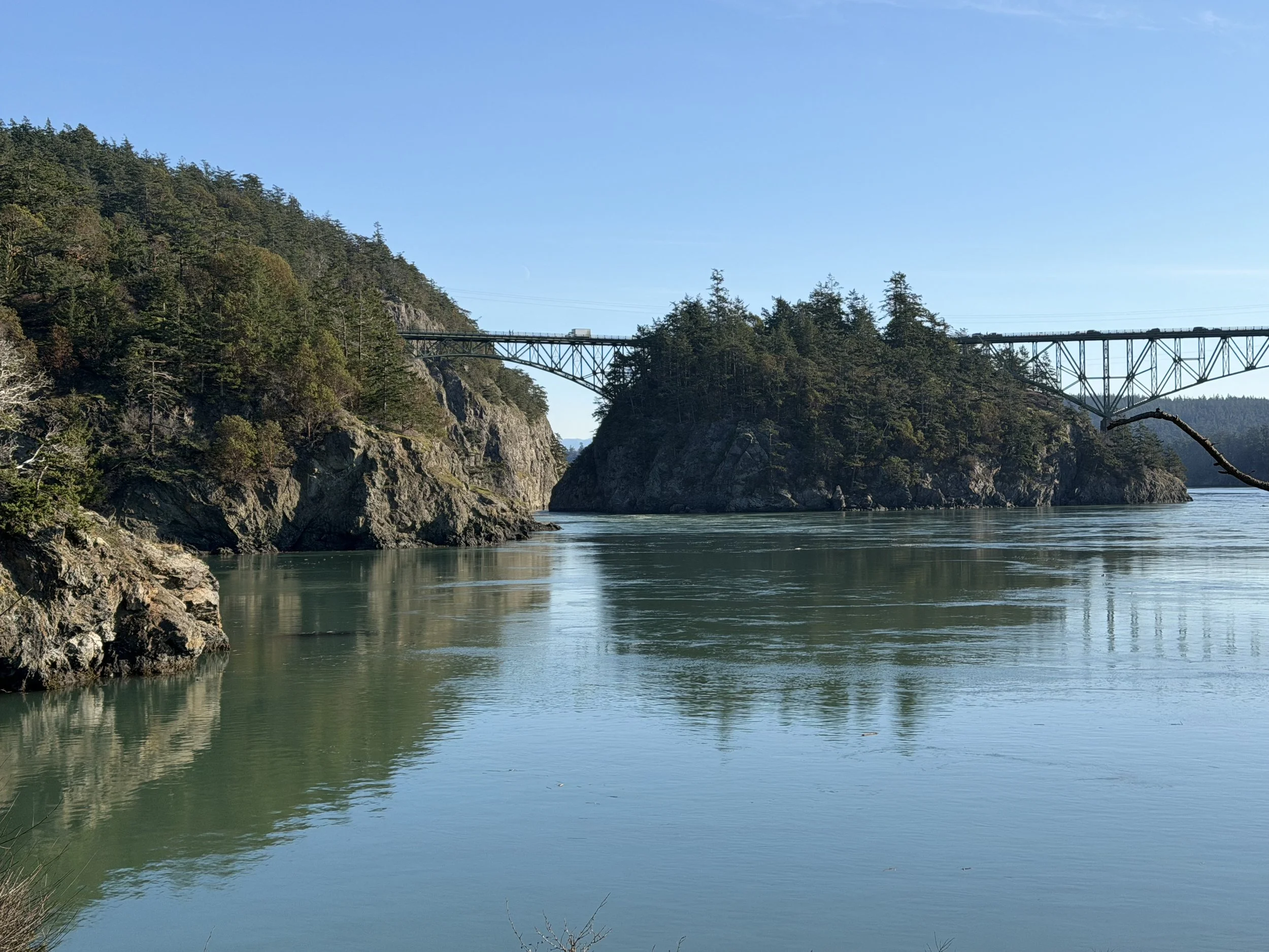 A scenic view of a river with calm water, surrounded by rocky, forested cliffs and a bridge spanning across the water in the distance under a clear blue sky.