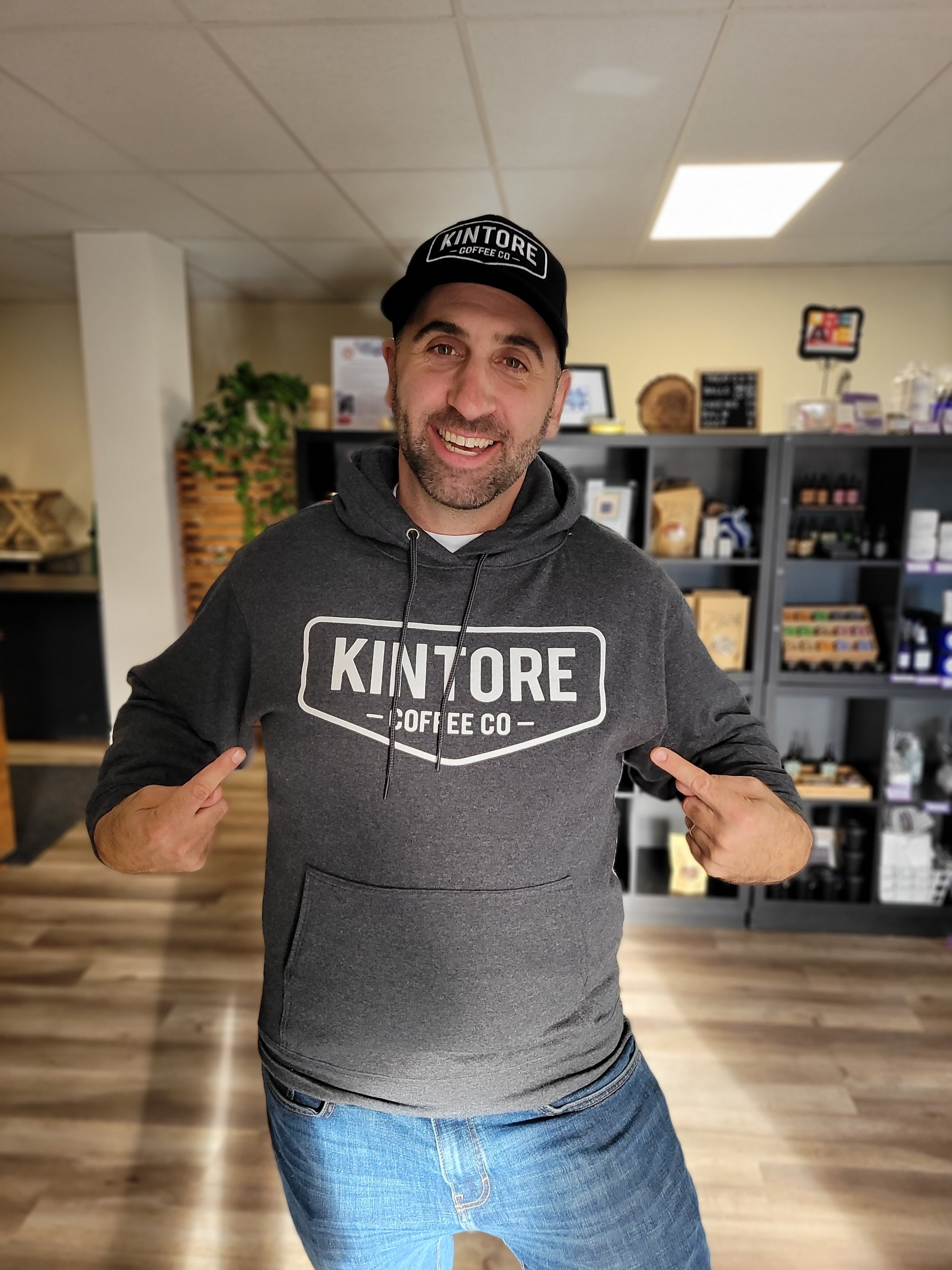 A person wearing a "Kintore Coffee Co" hoodie and cap, standing in a coffee shop with shelves of products behind them.