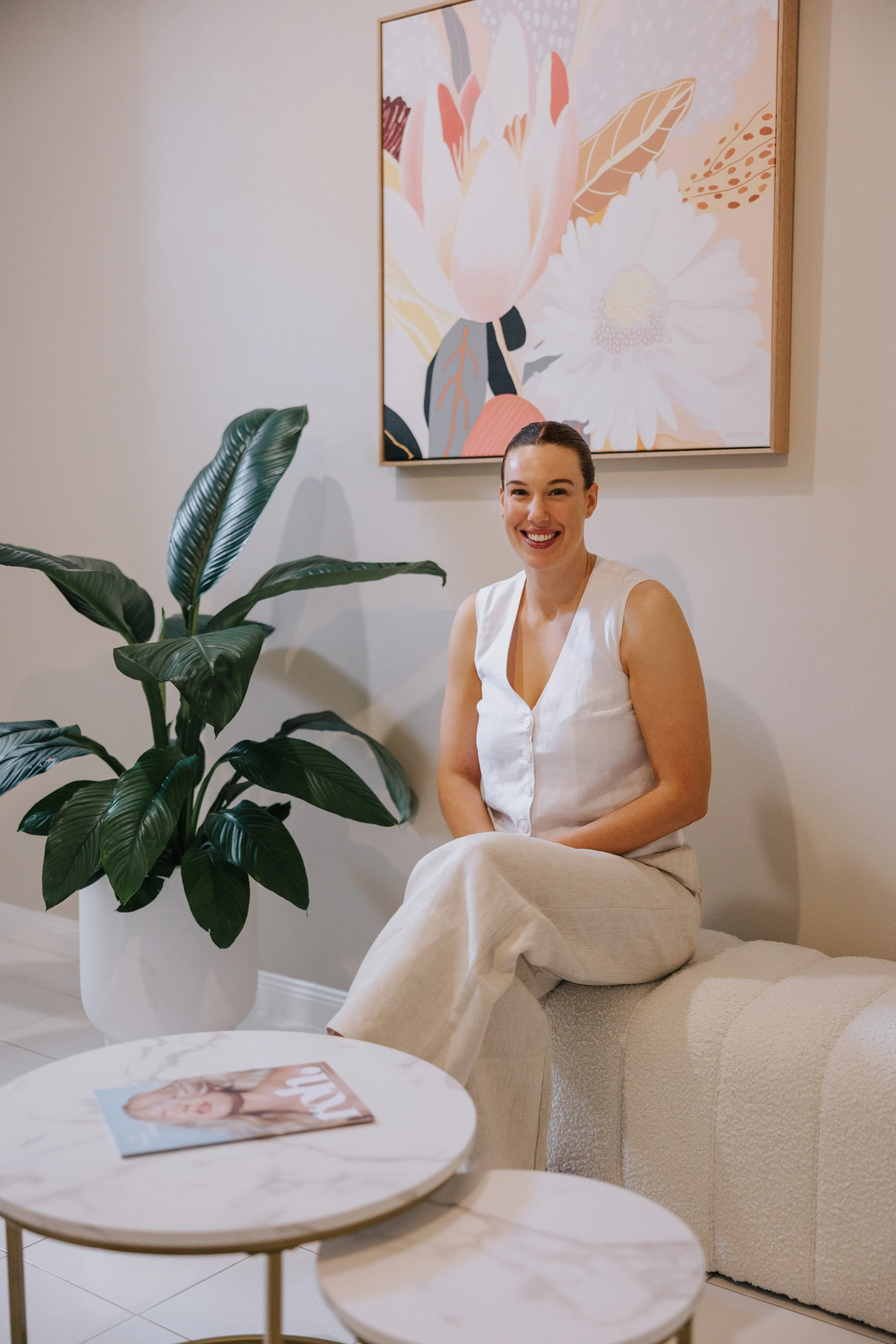 A woman in a white sleeveless top and light beige pants smiling while sitting on a cream-colored sofa in a modern living room with a large green plant and an abstract floral artwork on the wall behind her. There is also a round marble coffee table with a magazine on it.