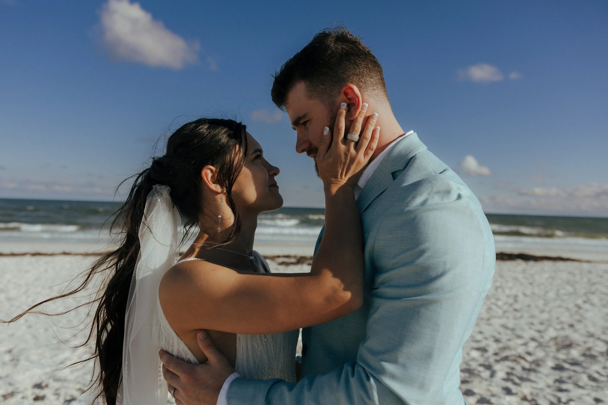A couple in wedding attire on the beach, holding each other and gazing into each other's eyes, with the ocean and sky in the background.