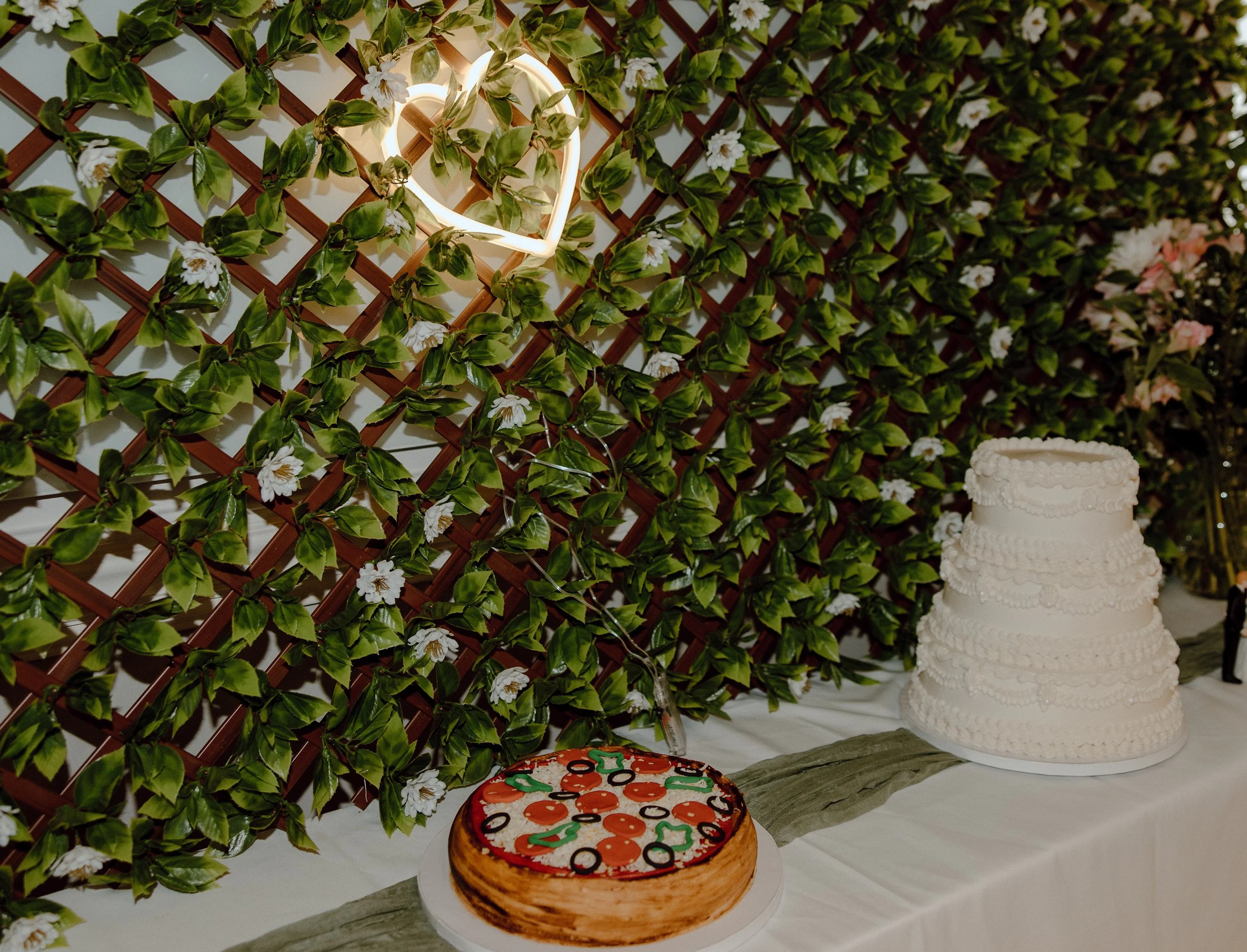 A table with a white tablecloth displaying a white tiered wedding cake and a pizza-shaped cake against a backdrop of a green leafy lattice wall decorated with white and pink flowers and a glowing heart-shaped light.