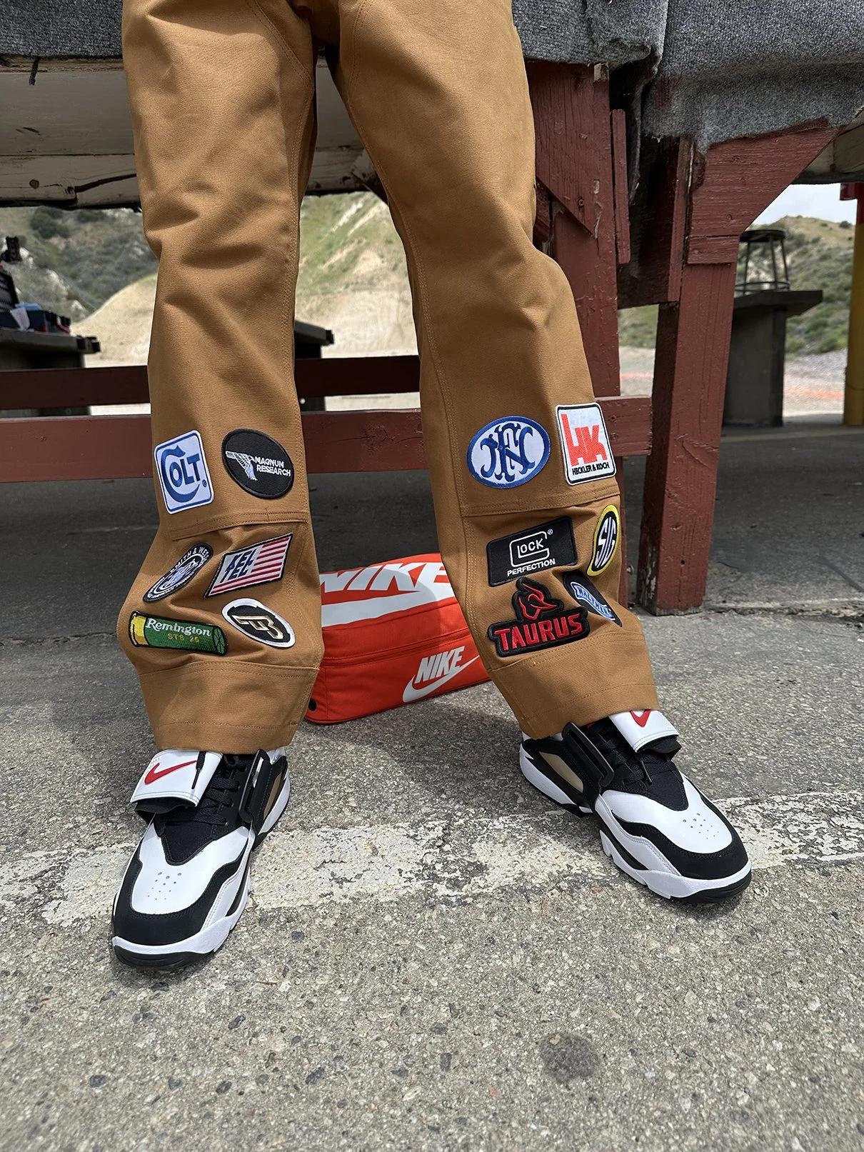 Person wearing tan pants with various patches standing on concrete ground, with Nike shoes, and a Nike bag in the background.