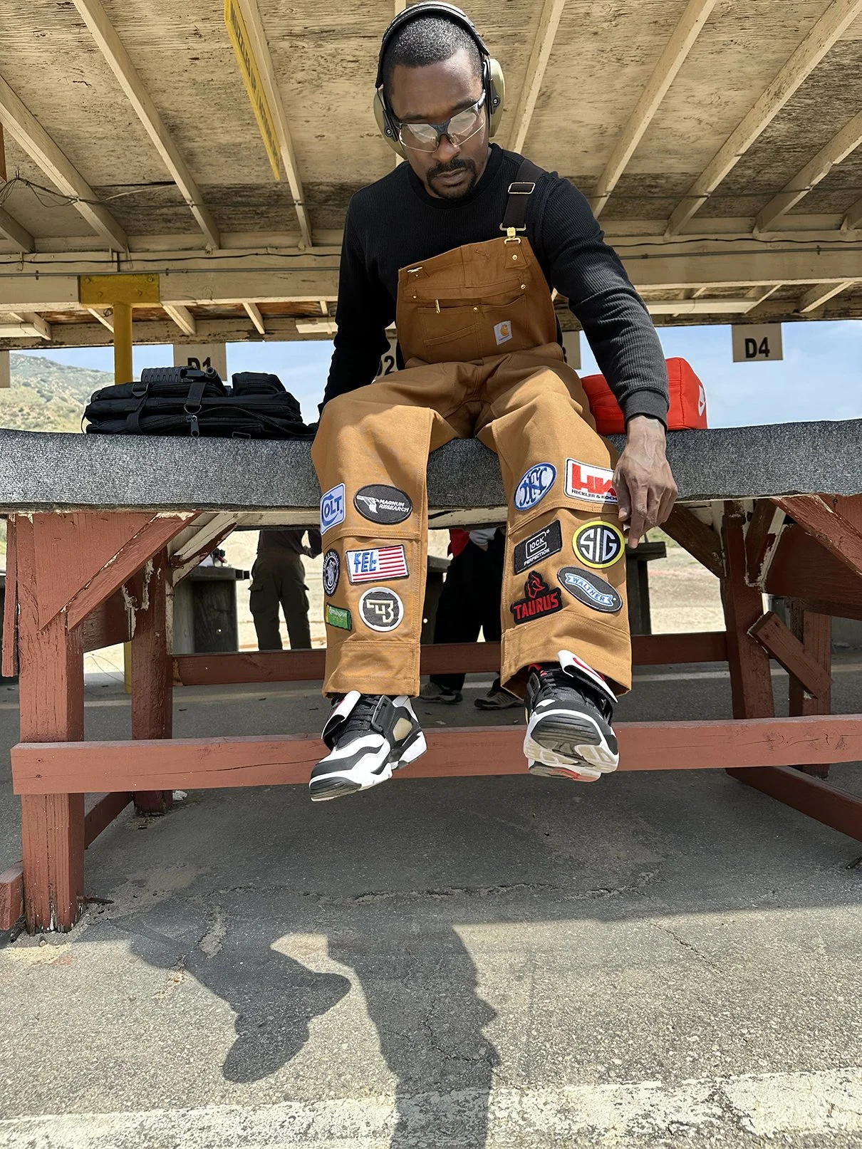 Clarence Nathaniel, Roses N' Guns founder, sitting on bleacher at shooting range, wearing safety goggles, headphones, and tactical gear with patches, with mountain and sky background.