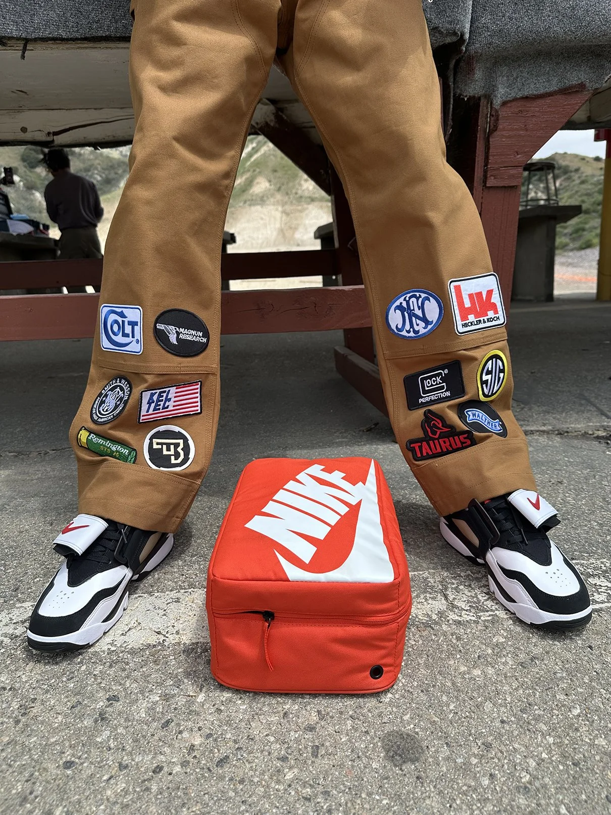 Clarence Nathaniel, founder of Roses N' Guns, wearing brown pants with numerous embroidered patch logos, white and black Nike sneakers, and standing on a concrete ground, with a red Nike-branded bag on the ground in front of him.