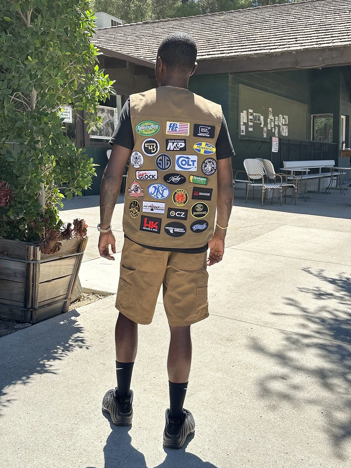 Clarence Nathaniel wearing tan Roses N' Guns vest decorated with numerous embroidered patches on the back, tan shorts, black socks, and black shoes, standing outside facing away from the camera near a building with green walls and outdoor seating.