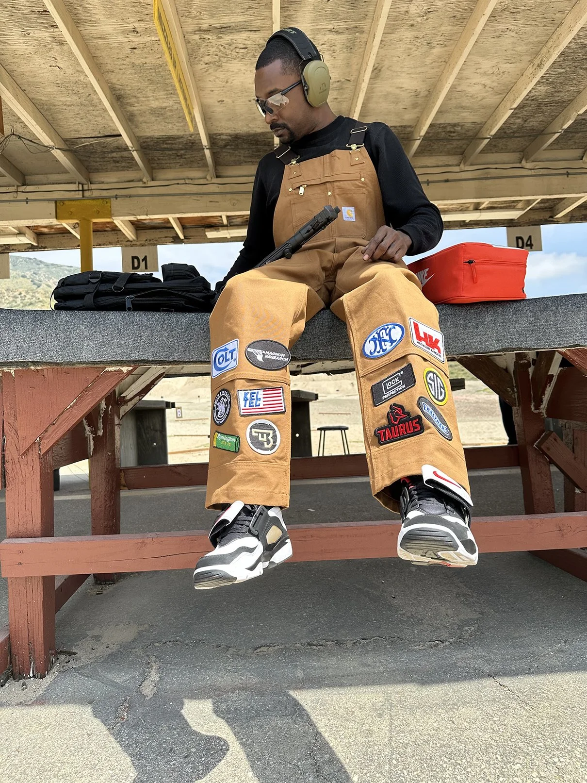 Clarence Nathaniel, founder of Roses N' Guns, on bench at an outdoor shooting range, wearing ear protection, sunglasses, a black shirt, brown shooting overalls with patch logos, holding a pistol, with shooting gear and a red first aid kit nearby.